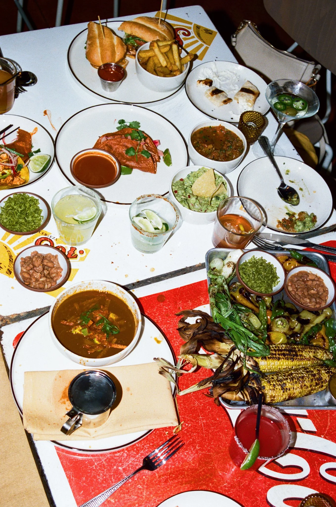 A table filled with Mexican food including grilled corn, tacos, guacamole, various salsas, soups, and drinks.