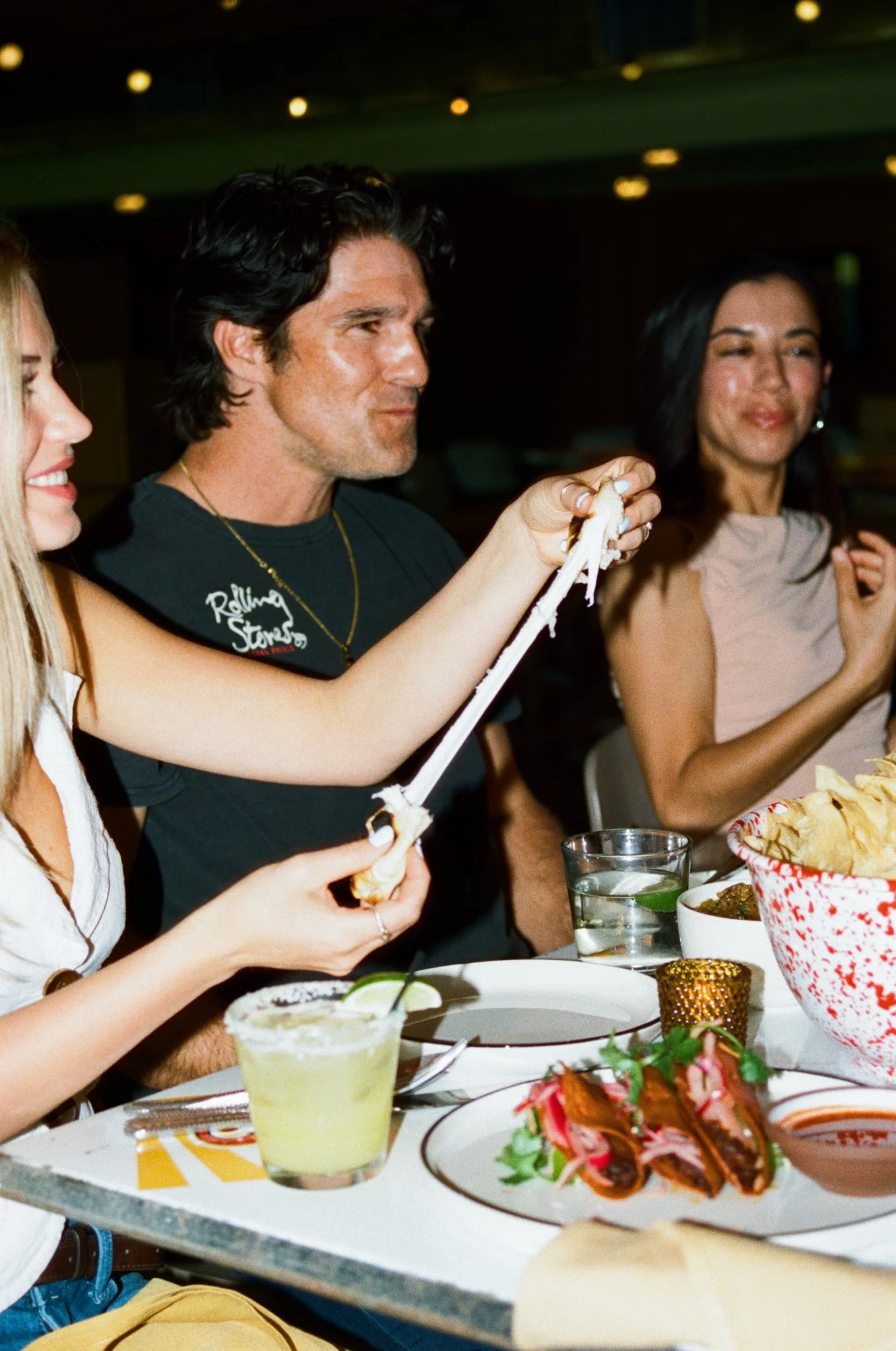Person stretching mozzarella cheese with hands at a dining table surrounded by food and drinks, including tacos, chips, and cocktails, with two other women smiling nearby.