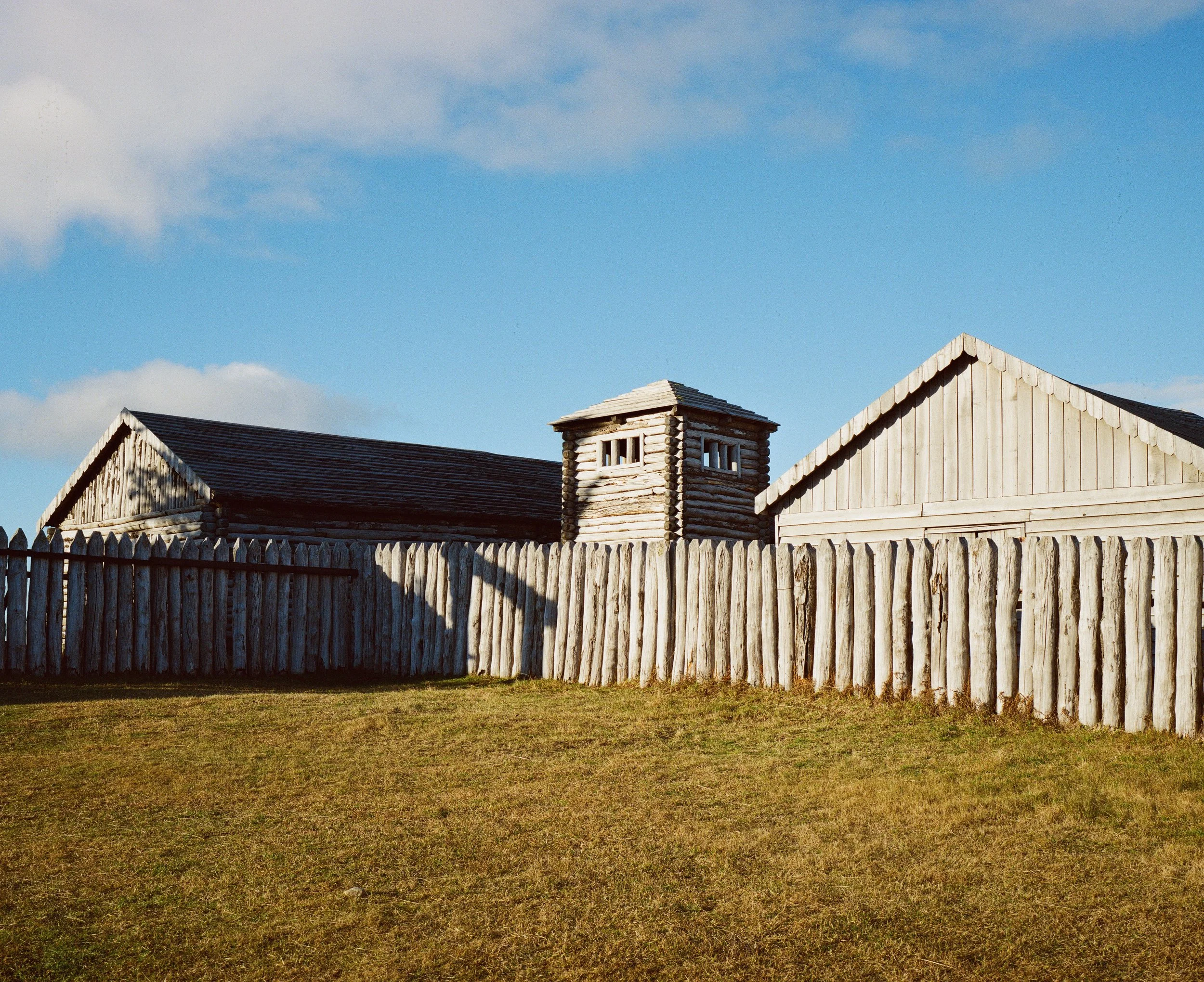 A wooden wagon wheel fence surrounds a grassy yard in front of two log-style buildings with sloped roofs, one with a small watchtower.