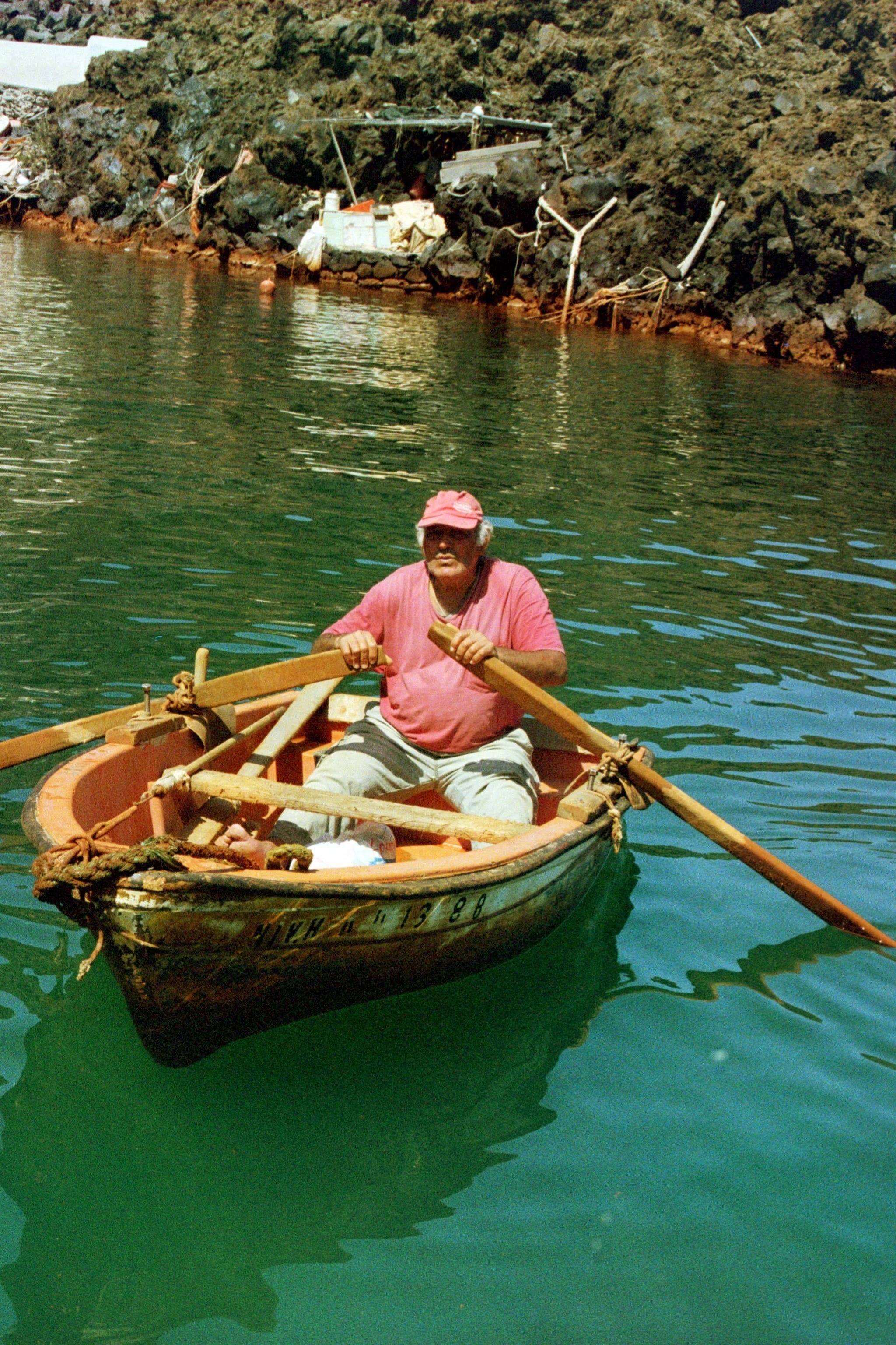 A man rowing a small boat on green water near a rocky shoreline.