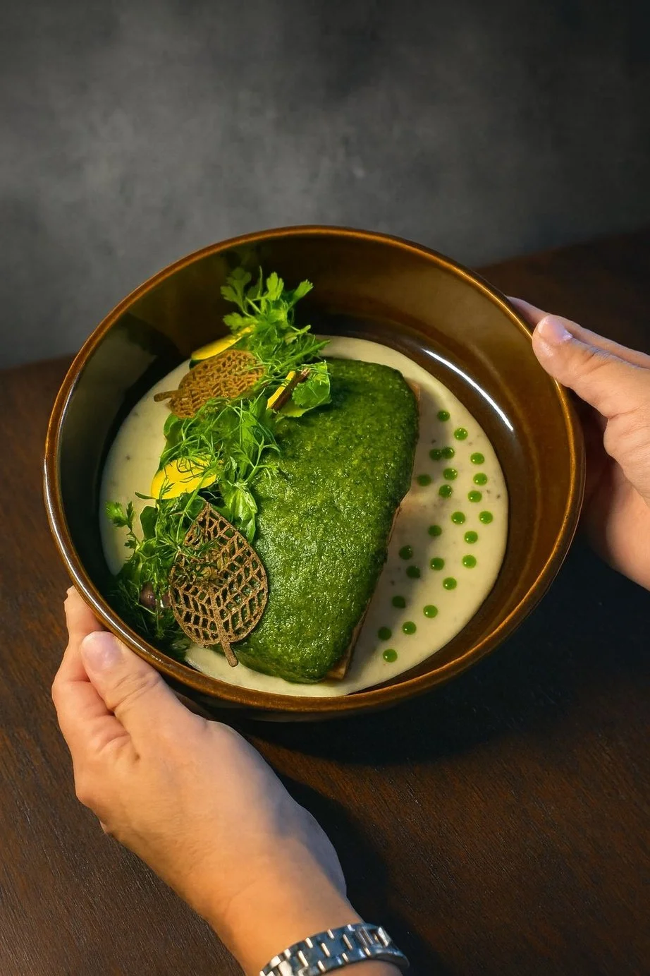 A ceramic bowl containing a large piece of green vegetable, garnished with herbs, decorative leaves, and small green sauce droplets, all on a creamy white sauce.
