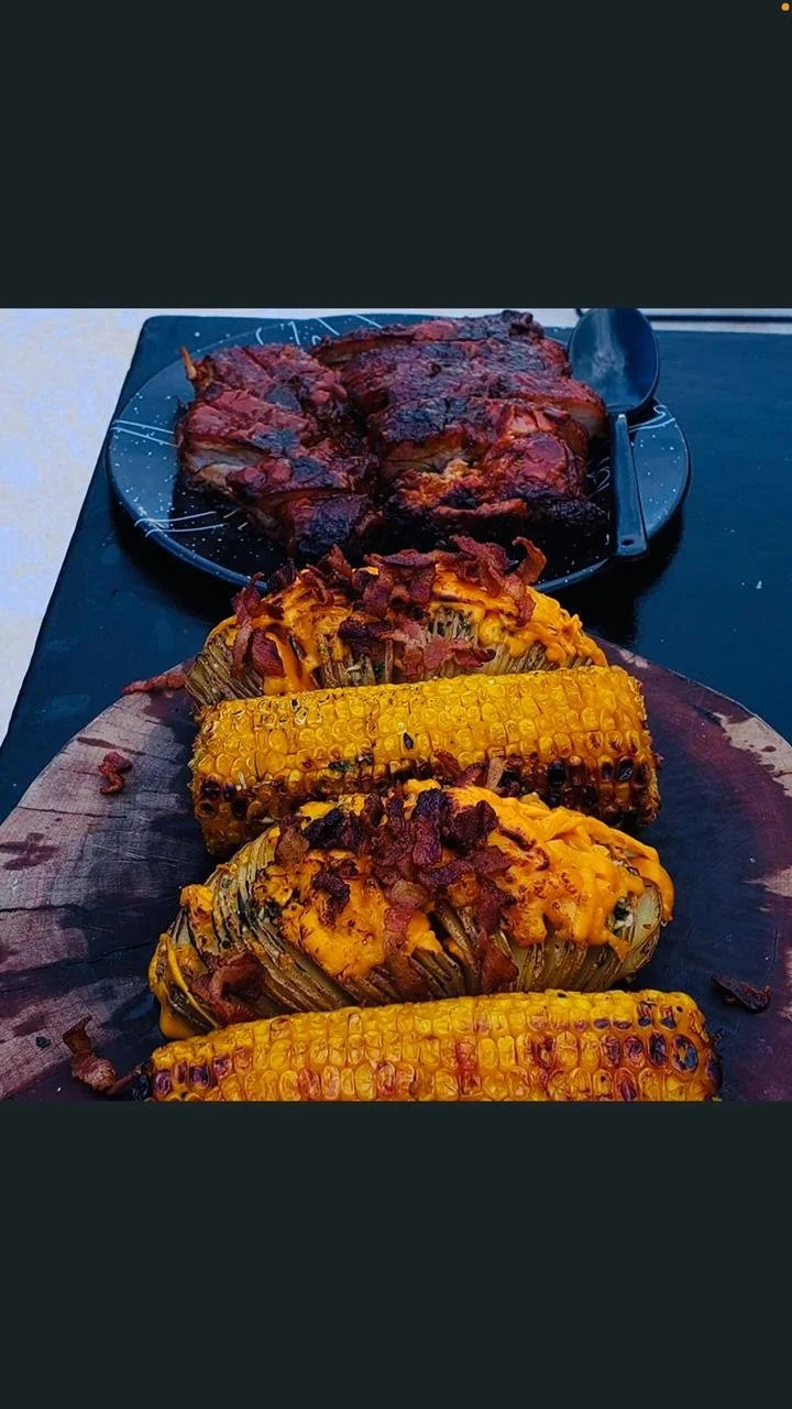 Grilled corn on a wooden cutting board topped with cheese and bacon, with a plate of barbecued ribs in the background.