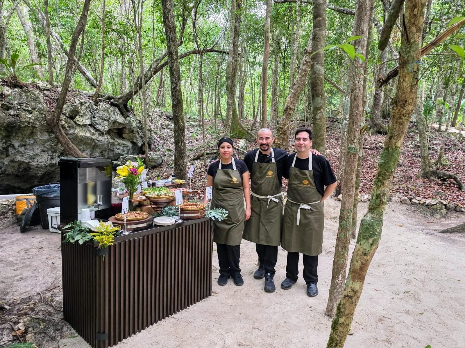 Three people standing behind a table with food and flowers in a wooded outdoor setting.