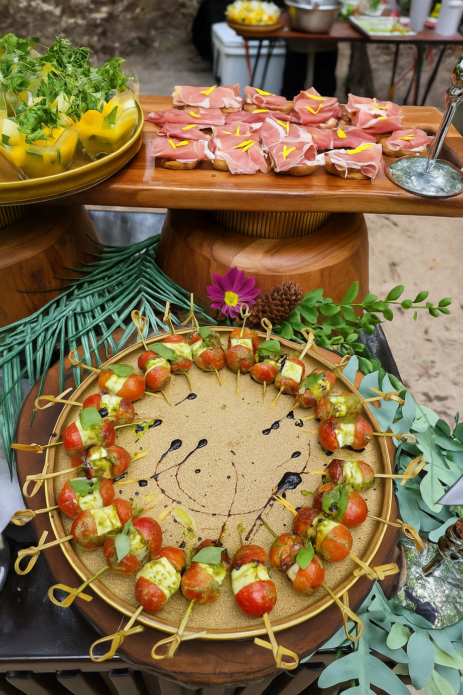 Assorted appetizers including cherry tomatoes with mozzarella and basil, prosciutto-wrapped bread slices with yellow flower petal garnish, and a bowl of salad with lettuce, yellow peppers, and other greens.