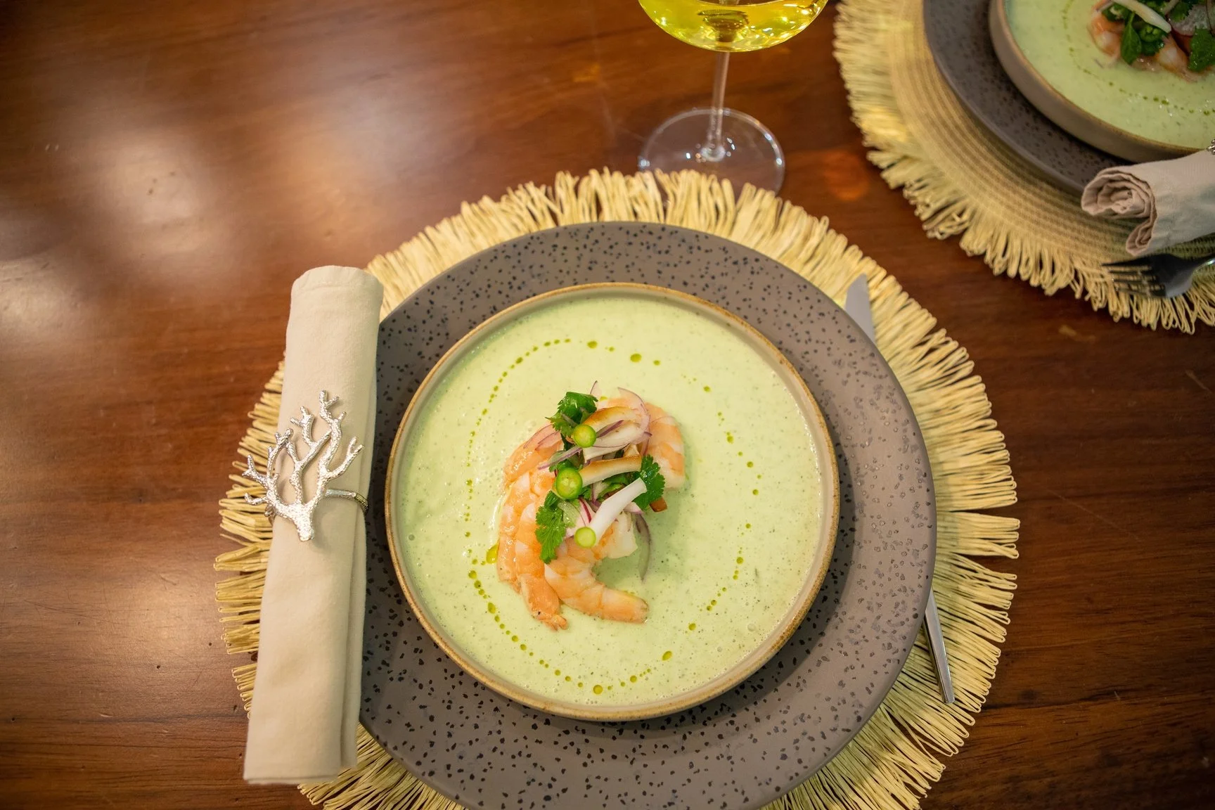 Bowls of green soup garnished with shrimp, sliced green onions, and fresh herbs, served on decorative plates with textured placemats and a glass of white wine.