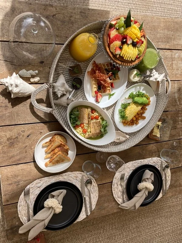 A wooden table set for a meal with a large silver basket of various dishes, including a fruit-topped cake, salads, bread, and beverages. There are two black plates with napkins and cutlery, some glasses, and a cleared glass dome on the left.