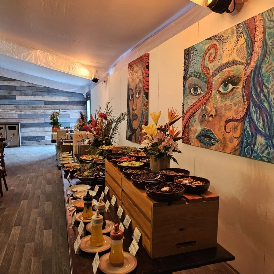 A buffet setup with various bowls of salads and toppings on a wooden table, decorated with flowers, inside a room with artistic portraits of women on the wall.