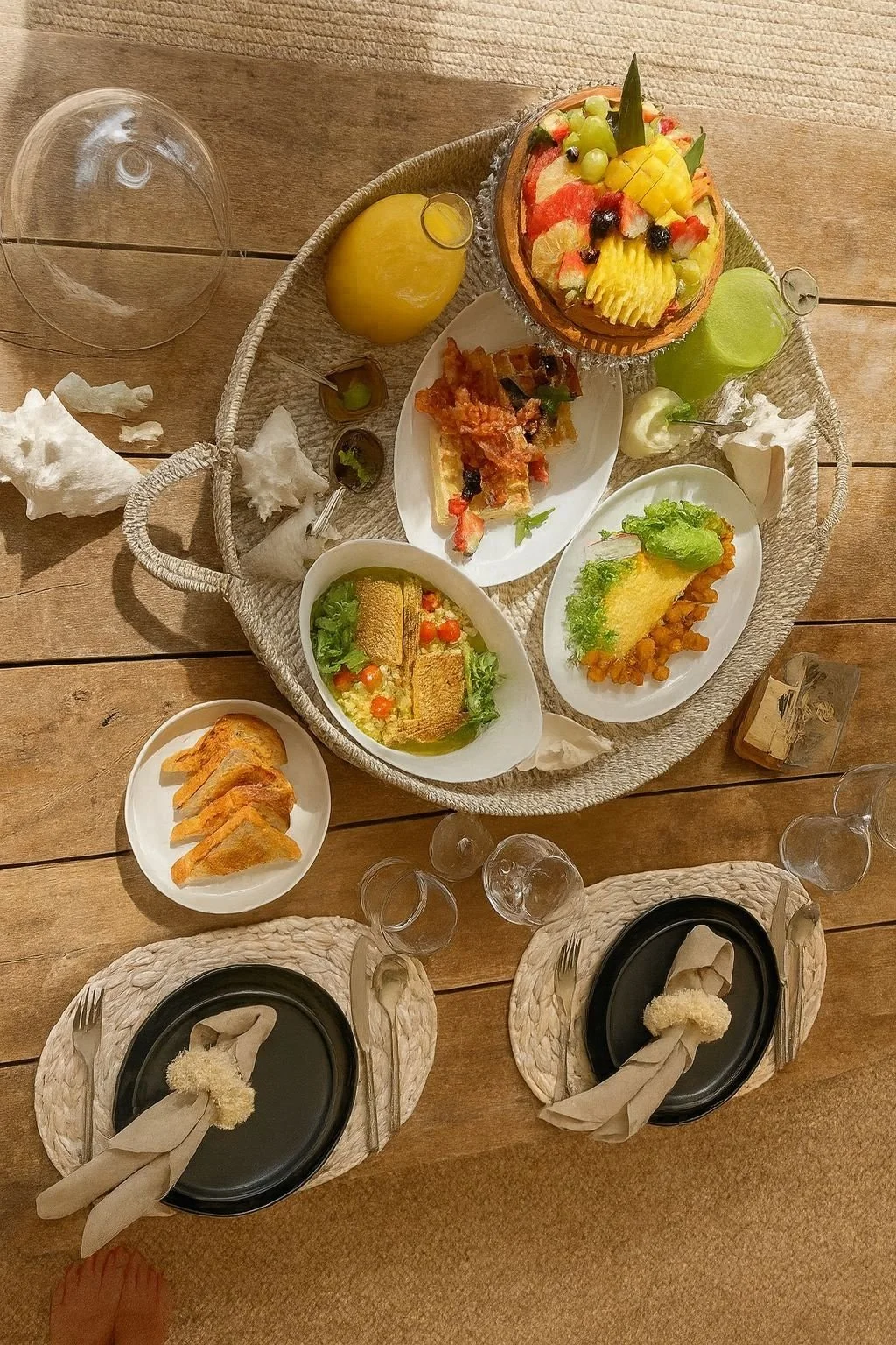 Overhead view of a wooden table with a woven tray holding various desserts and small plates, including a fruit-topped cake, slices of pie, and other sweet treats, with plates, glasses, napkins, and utensils arranged around it.