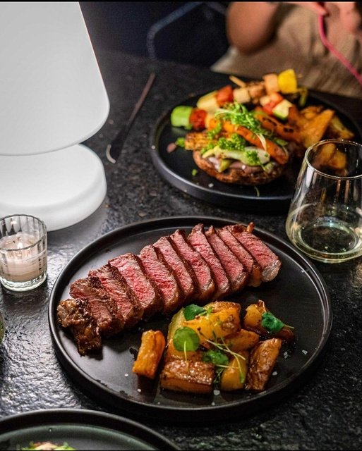 Sliced medium-rare steak with roasted vegetables on a black plate, with a glass of white wine nearby.