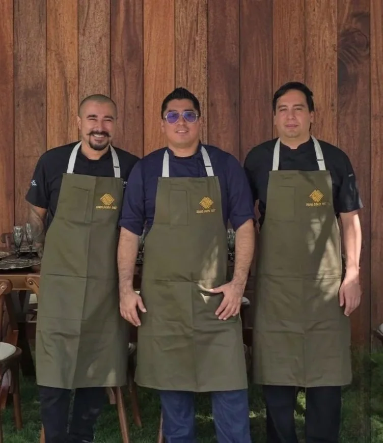 Three men wearing black chef uniforms and green aprons standing in front of a wooden wall, smiling.