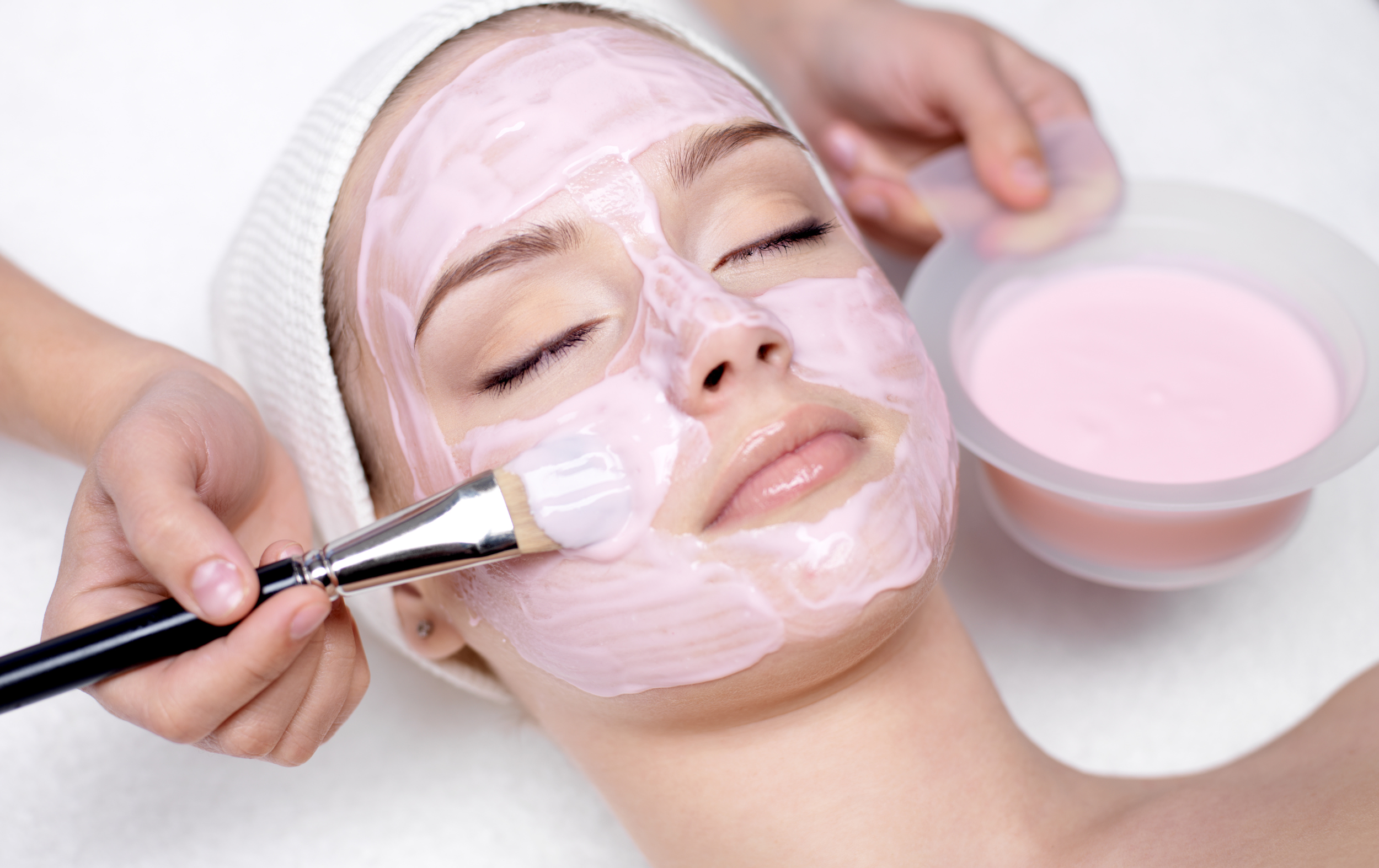 A woman receiving a facial treatment with pink cream being applied to her face during a spa session.