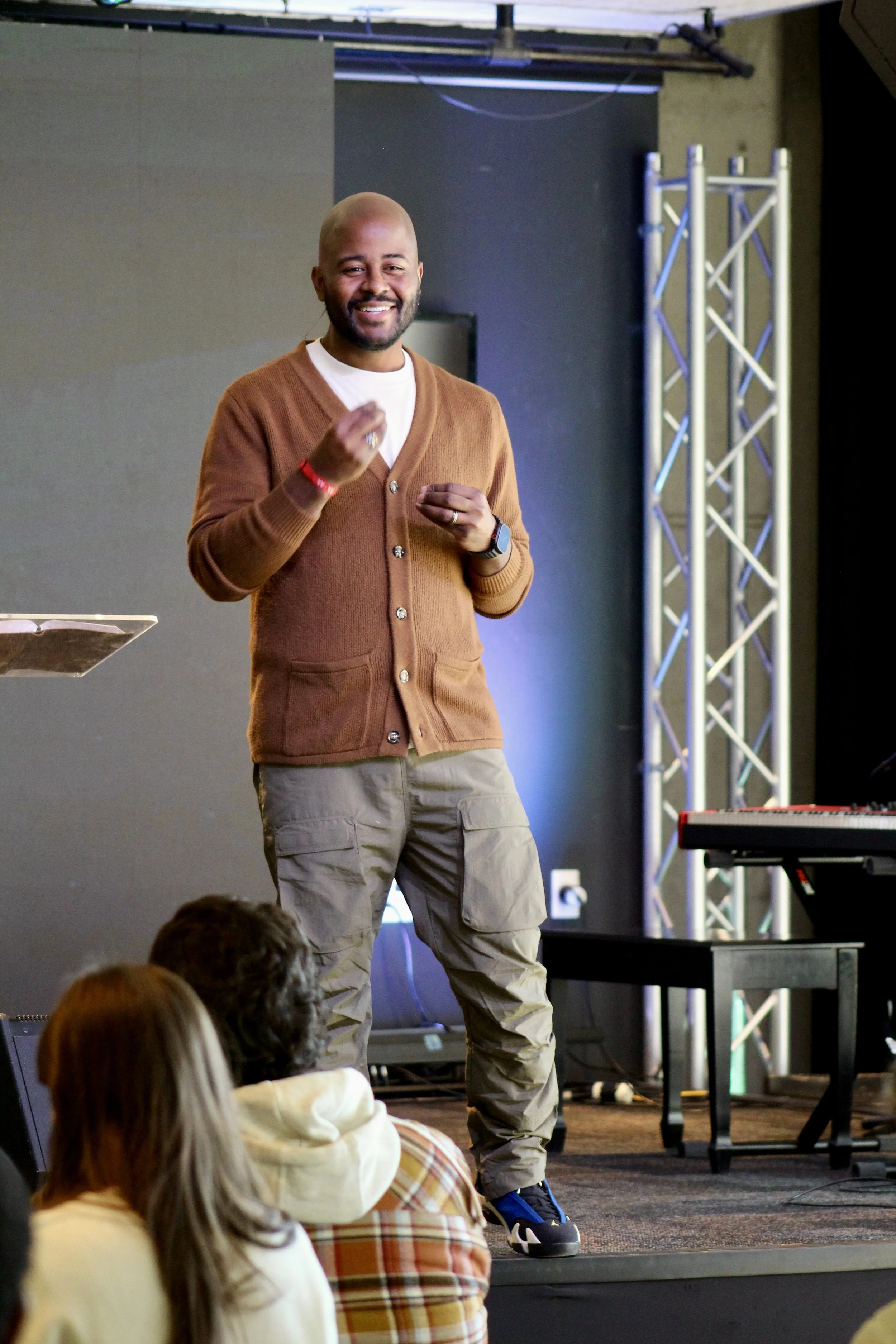 A man standing on stage smiling and gesturing with his hands, wearing a brown cardigan, khaki pants, and sneakers, with an audience seated in front of him.