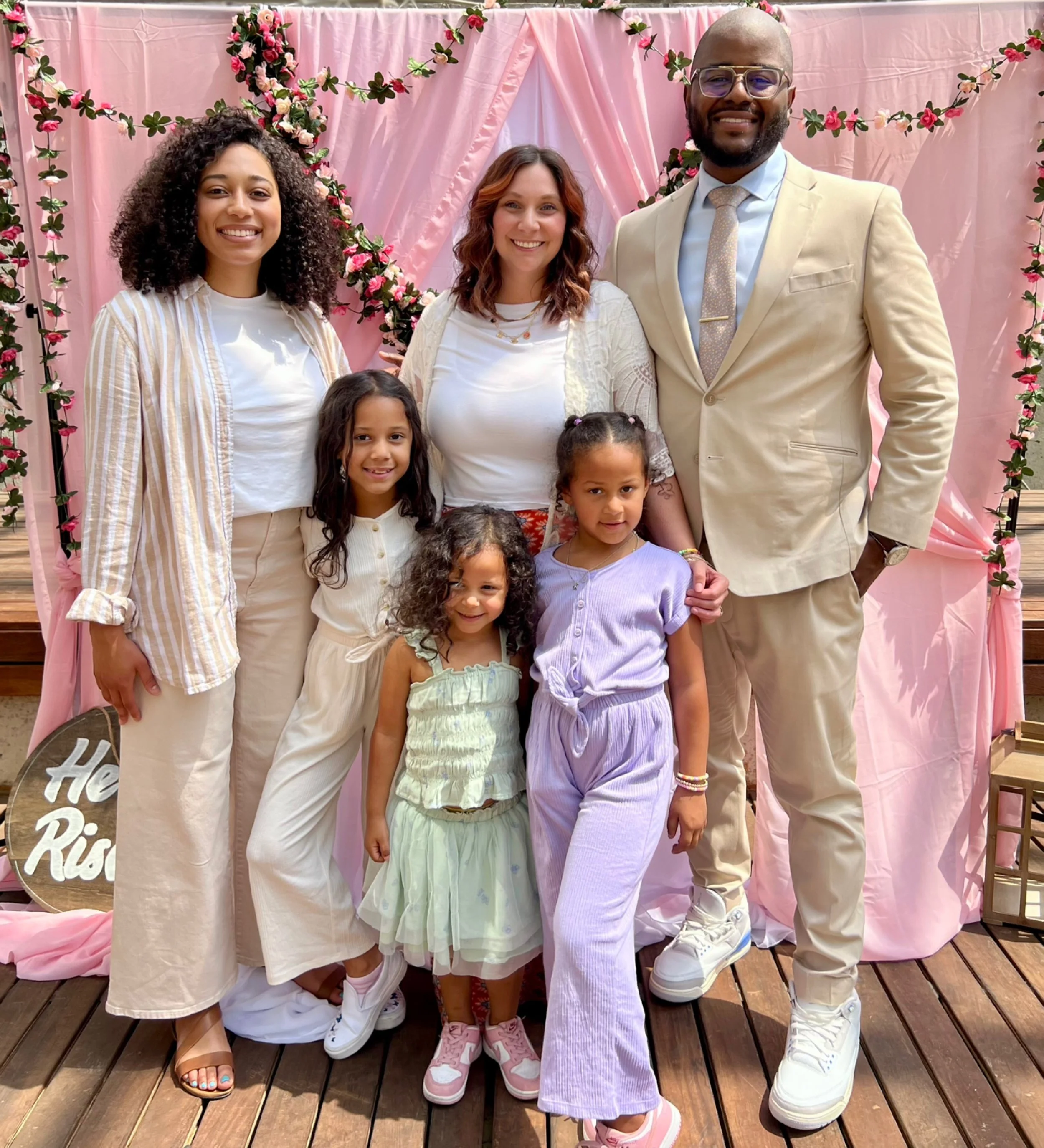 Family of seven posing in front of a pink floral decorated backdrop at a celebration, including adults and children dressed in light, pastel, and neutral colors.