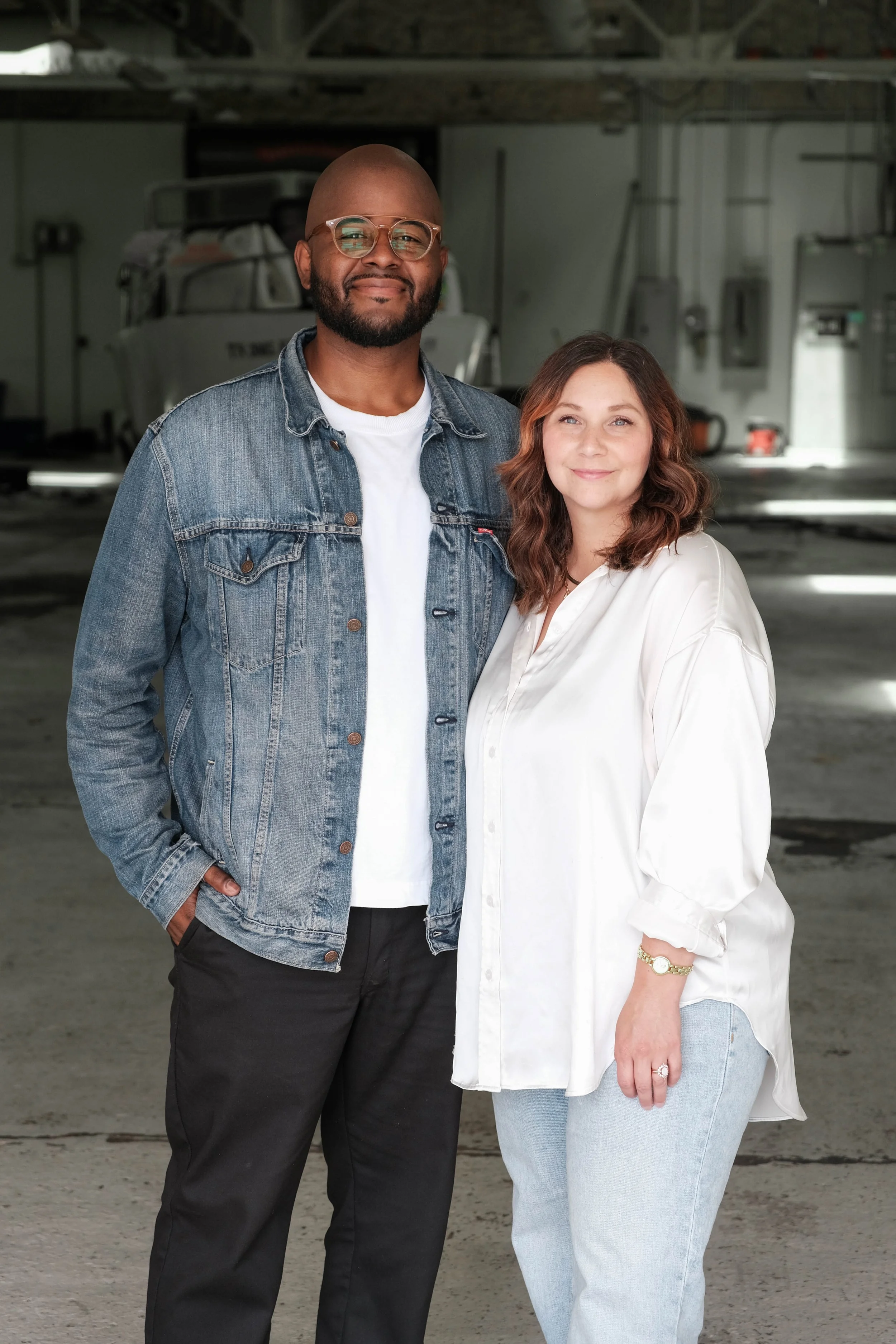 A man and woman standing side by side in a garage or warehouse, smiling at the camera.