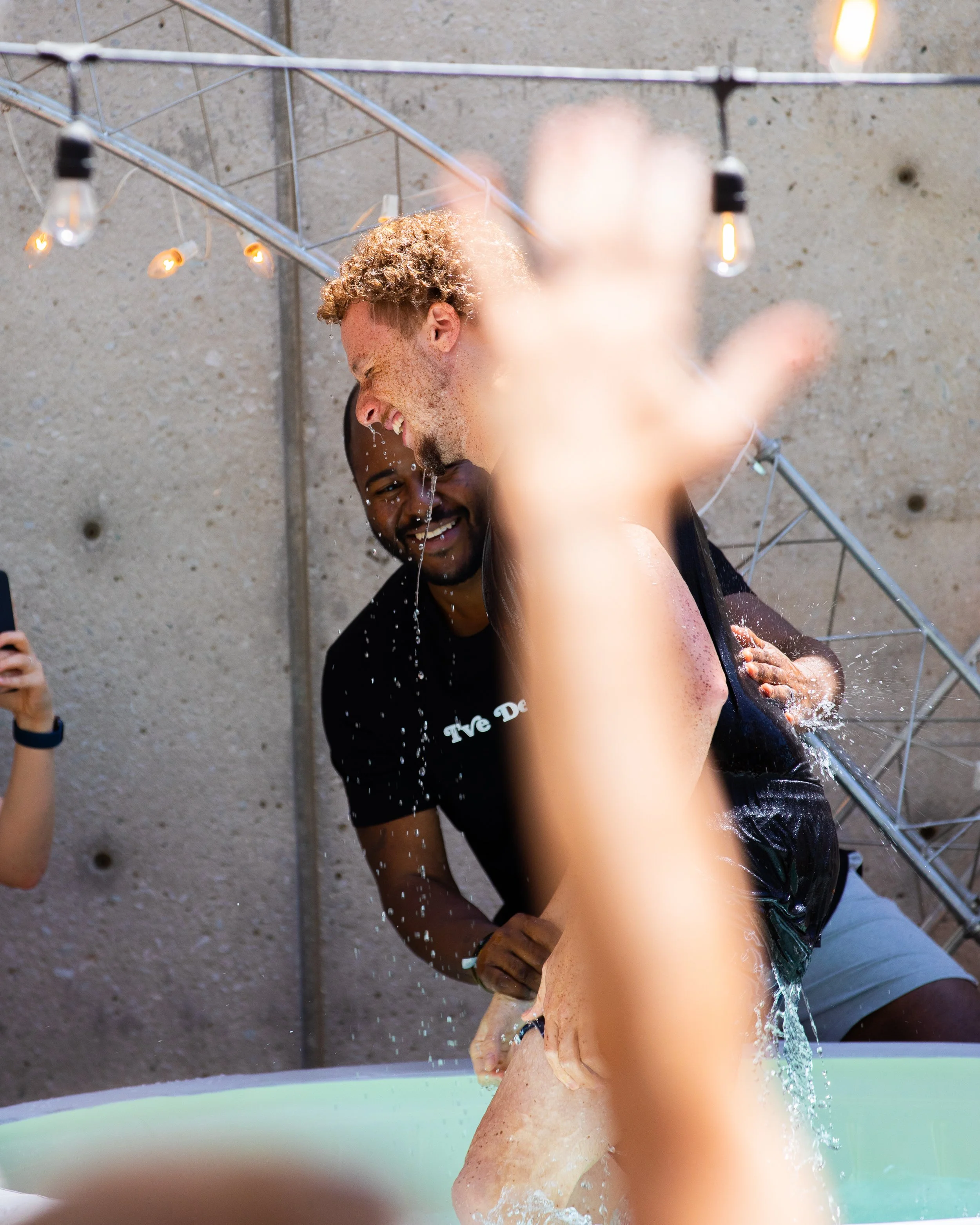 Two men laughing and enjoying a water activity outdoors, with one man splashing water on the other while they are by a concrete wall, decorated with string lights.