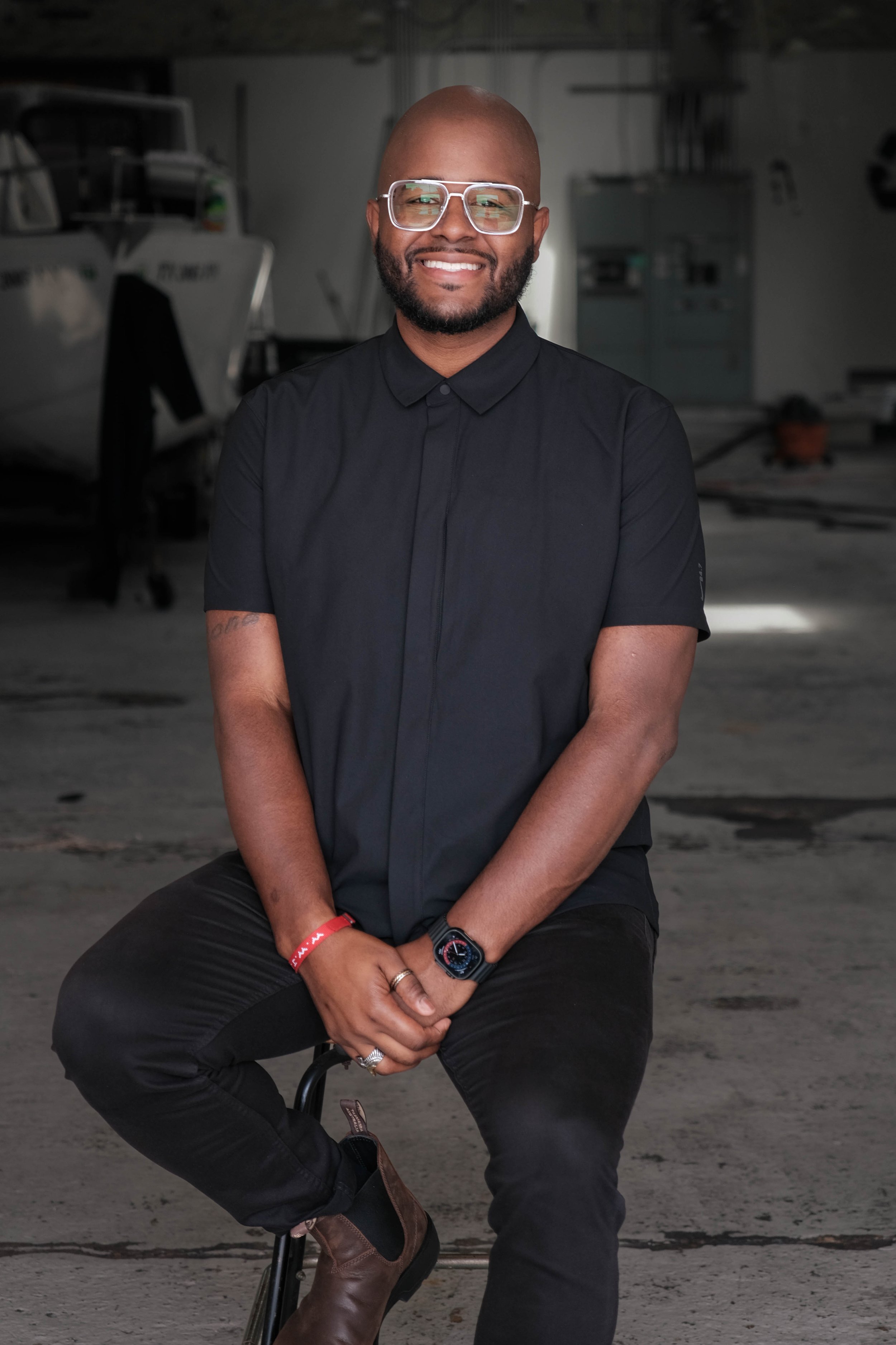A smiling man with glasses, a beard, and a bald head sitting on a stool in a garage or workshop with boats in the background, wearing a black shirt and black pants.