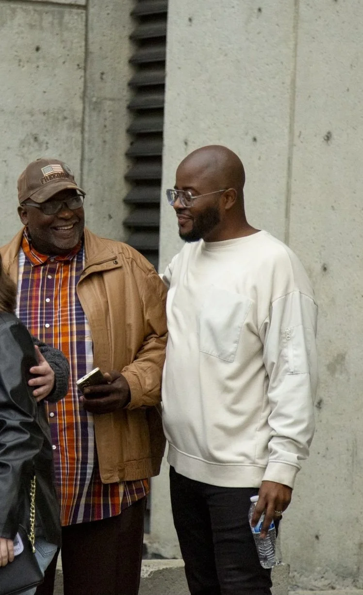 Two men smiling and talking outside, one wearing glasses, a brown jacket, and a plaid shirt, holding a phone, the other in a white sweatshirt, glasses, and holding a water bottle.