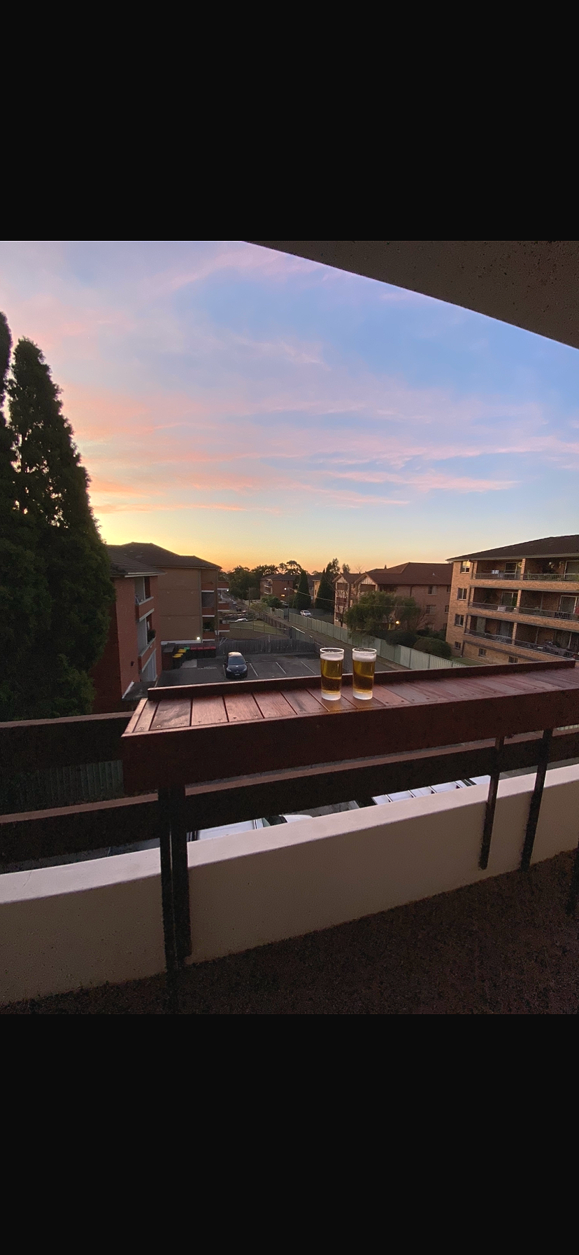 Two glasses of beer on a balcony railing overlooking a residential area at sunset with pastel pink and blue sky.