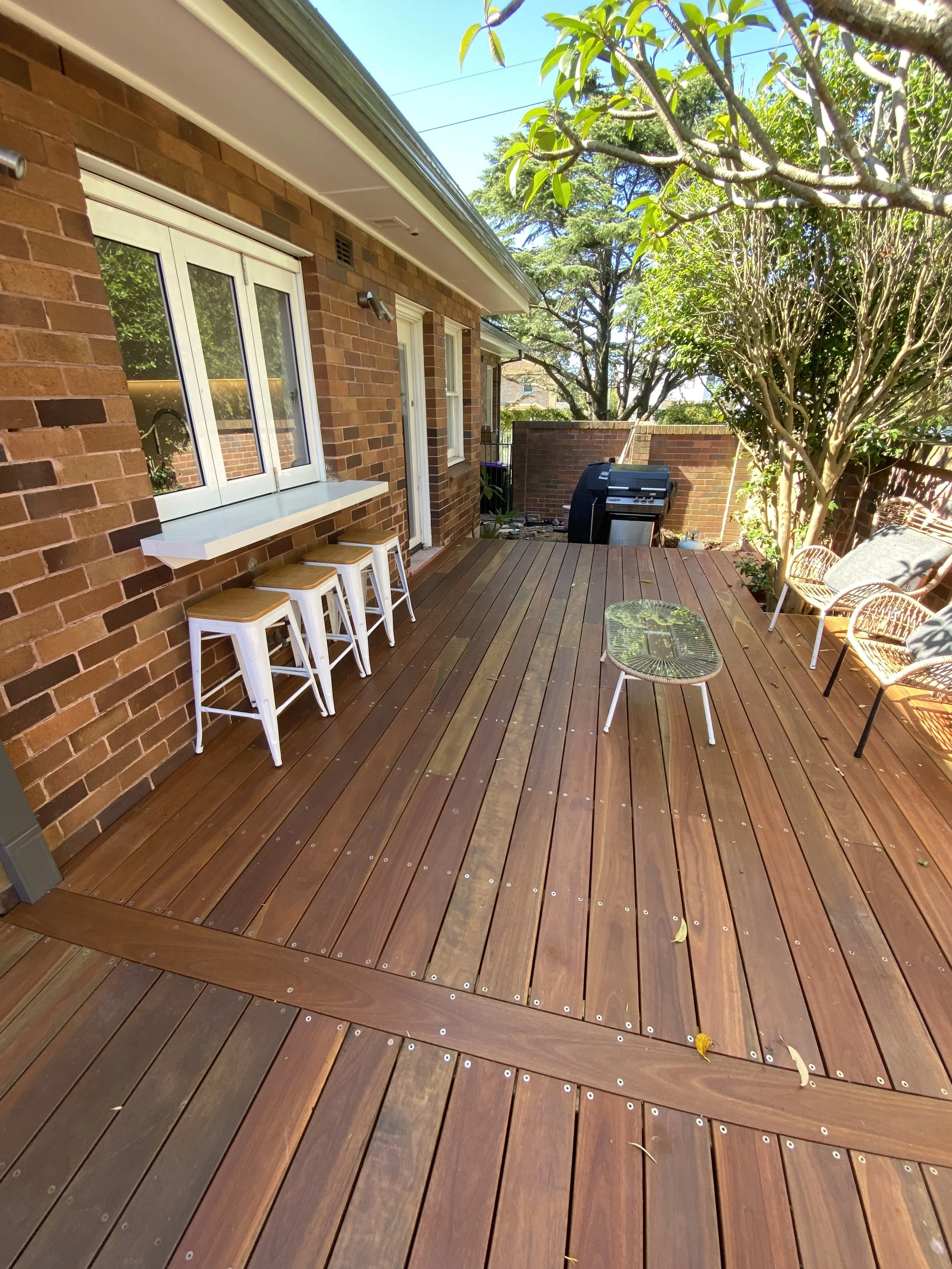 A spacious outdoor wooden deck featuring bar stools, a small table, and wicker chairs with cushions, surrounded by lush green trees and a brick wall at the back, with a barbecue grill nearby.