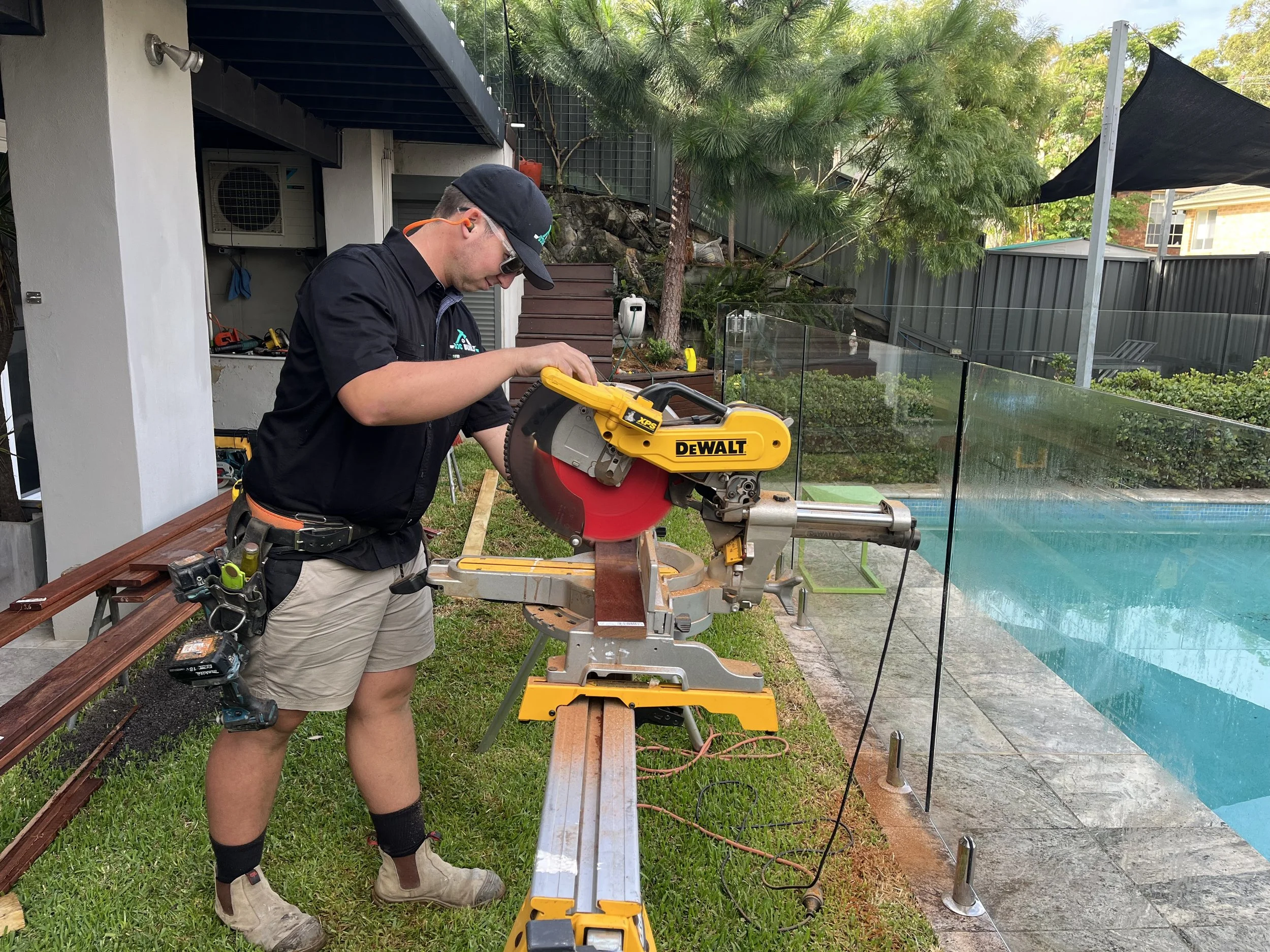 A man wearing safety glasses, a black cap, a black shirt, and shorts operates a yellow DeWalt miter saw on a sawhorse outdoors next to a swimming pool, cutting a piece of wood.