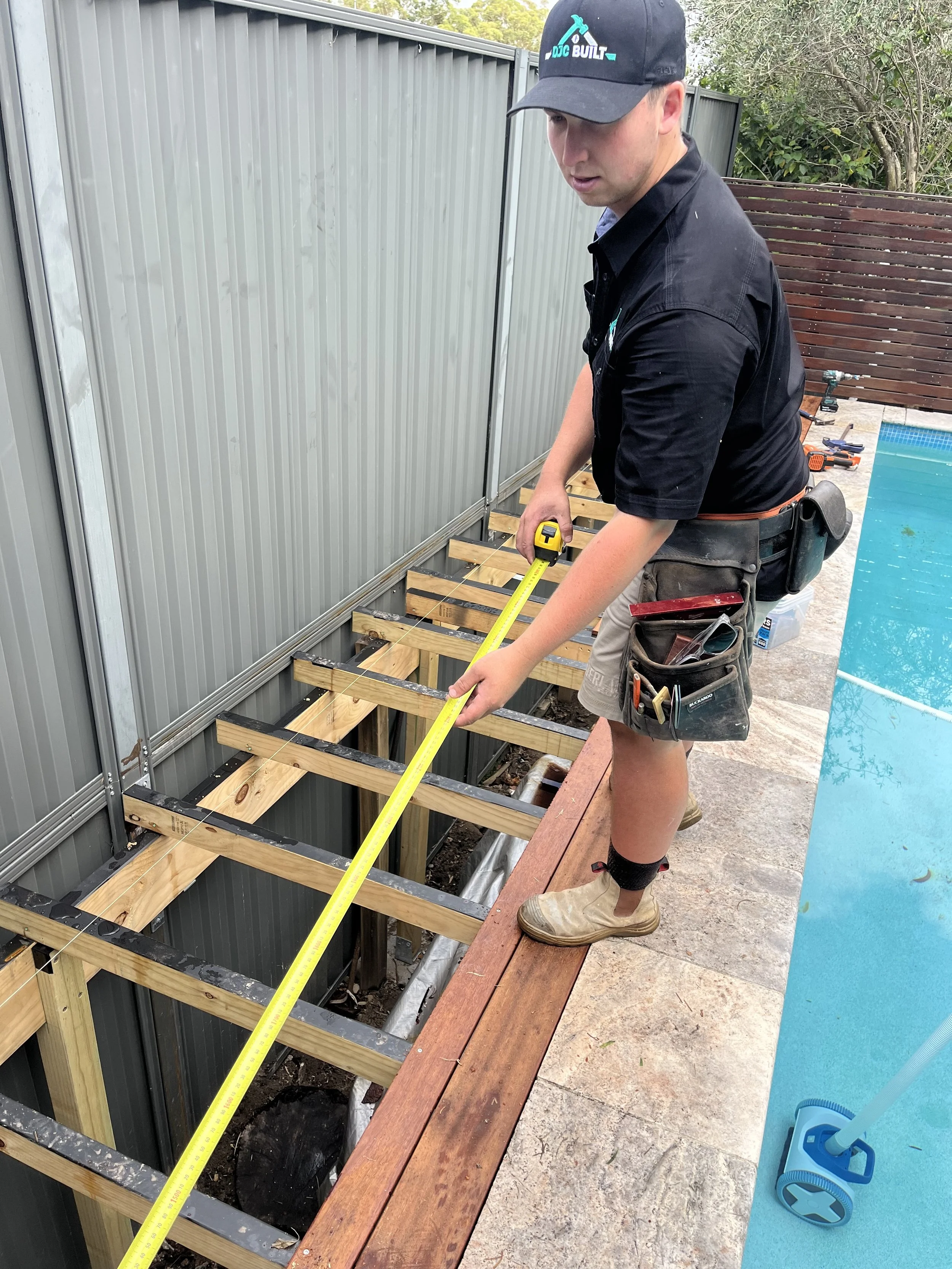 A man standing on a wooden deck edge using a tape measure to measure the distance between the deck and the ground, with a partially built wooden deck structure, tools, and a swimming pool in the background.