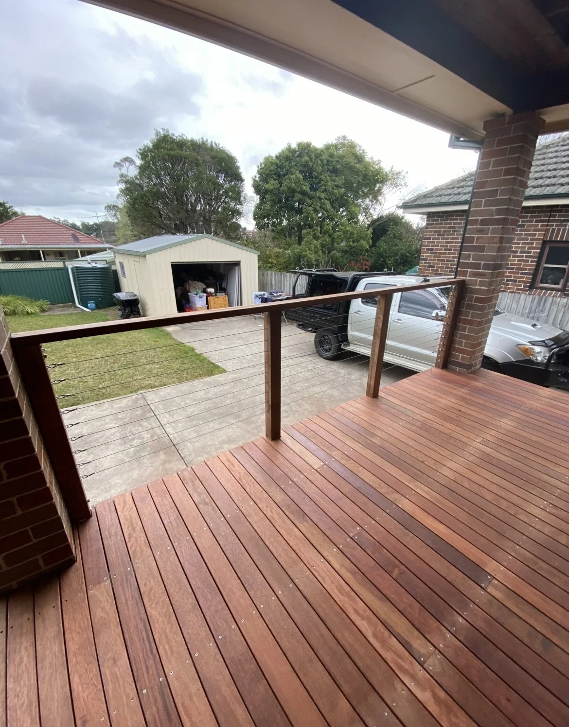 View from a new porch deck overlooking a backyard with trees, a small shed, a white truck, and a silver car parked on a concrete driveway.