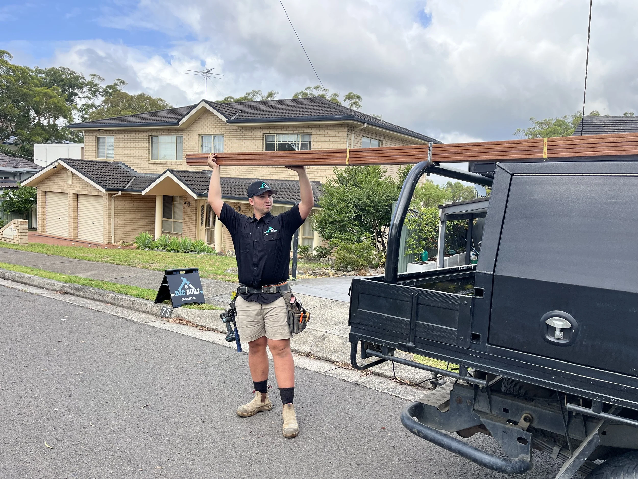 A worker carrying wooden planks on his shoulder standing near a black utility vehicle on a residential street.