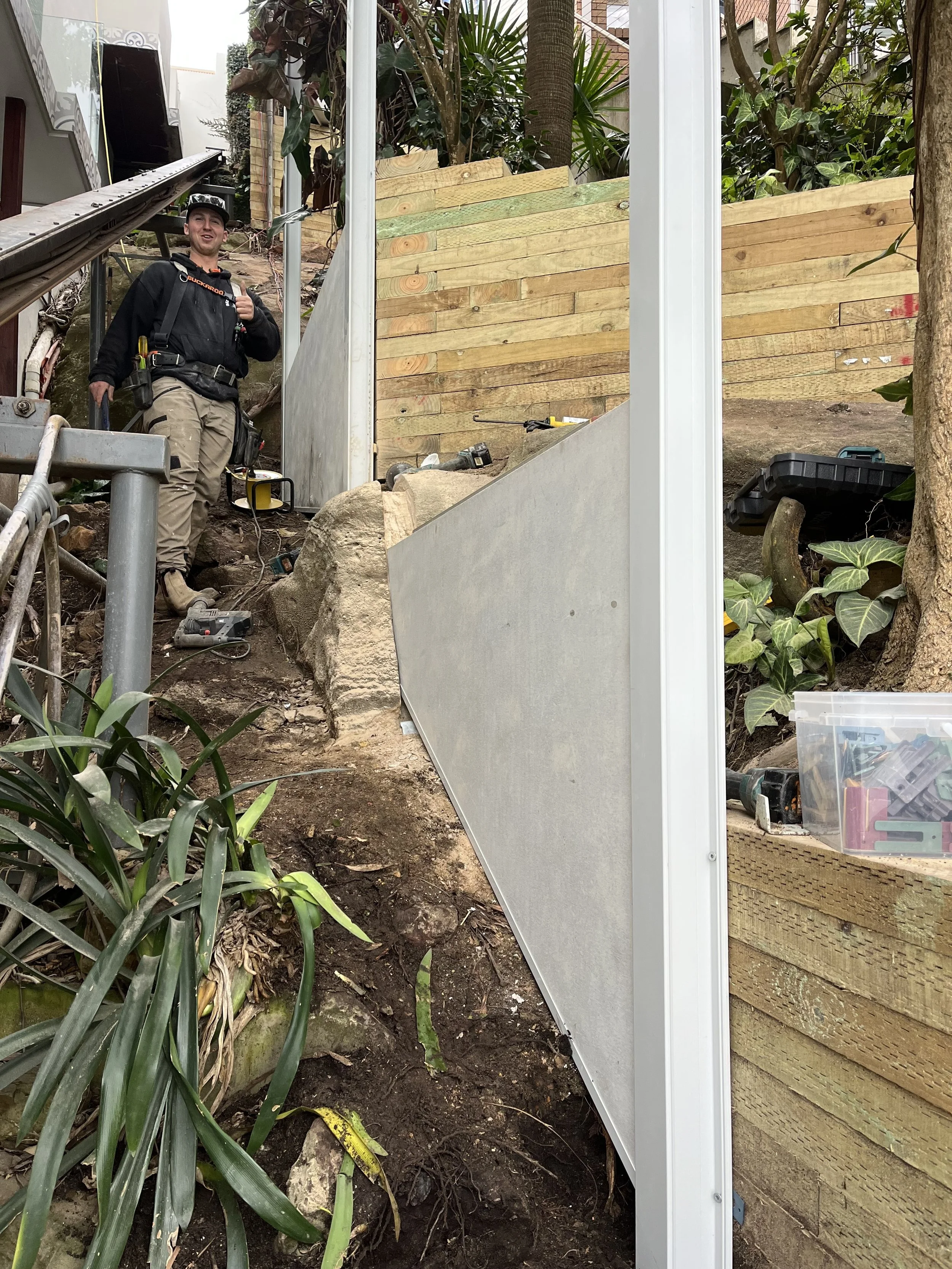 A man in construction gear standing on uneven dirt ground, smiling and giving a thumbs-up, with tools and construction materials around, next to a partially built wooden fence and garden plants.