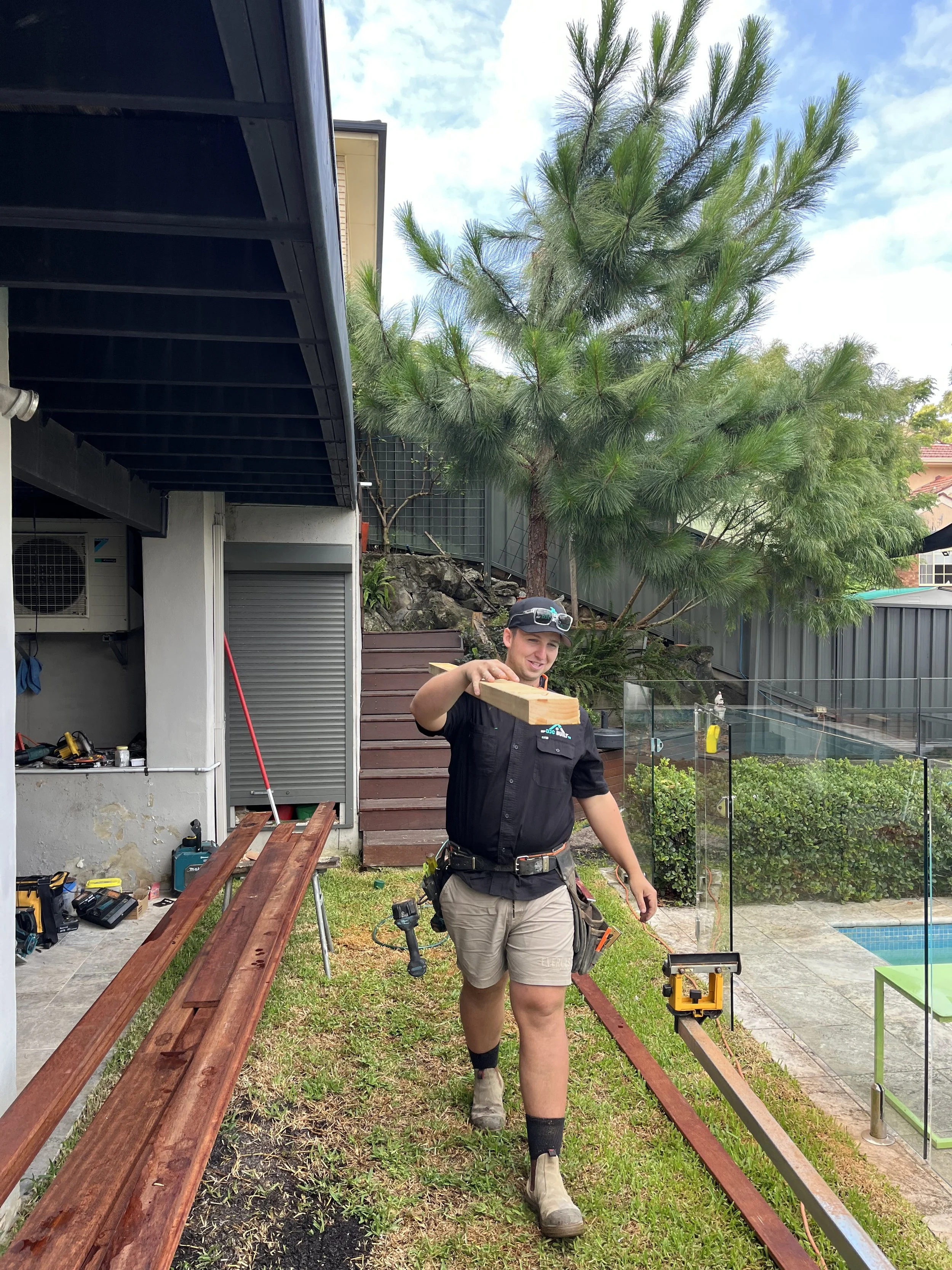 A person working outdoors next to a house, holding a piece of wood, with tools and construction equipment nearby, and a glass fence with greenery in the background.