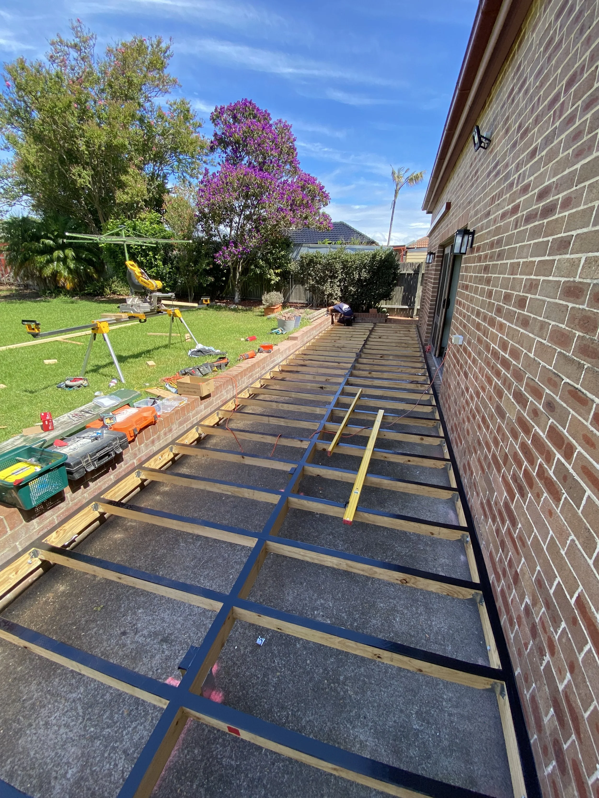 backyard construction site with wooden framework and tools for a deck or patio project, adjacent to a brick house, with a grassy yard, trees, and blue sky in the background.