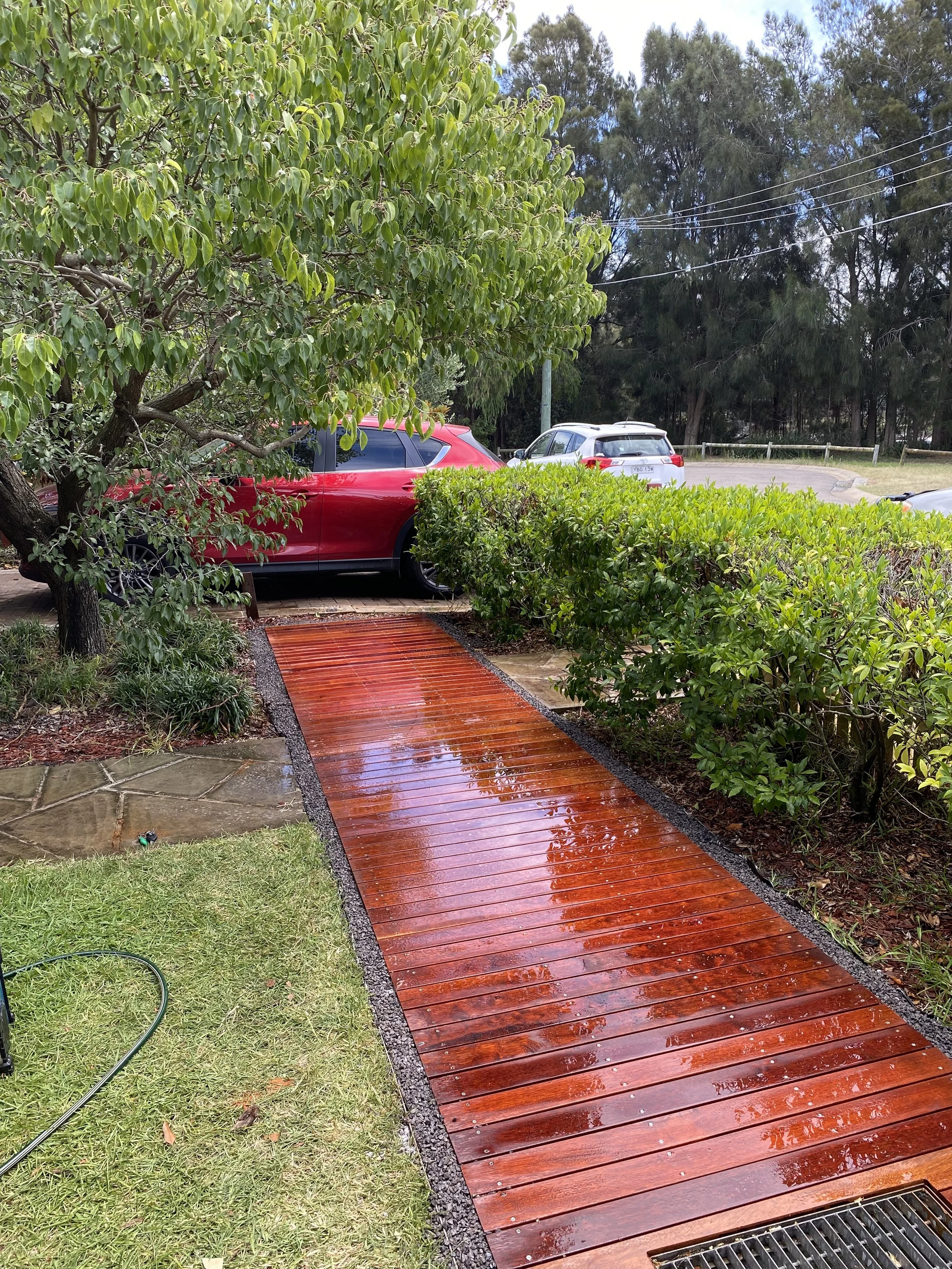 Wet wooden pathway leading to parked cars partially obscured by trees and bushes, with grassy area and nearby drain.