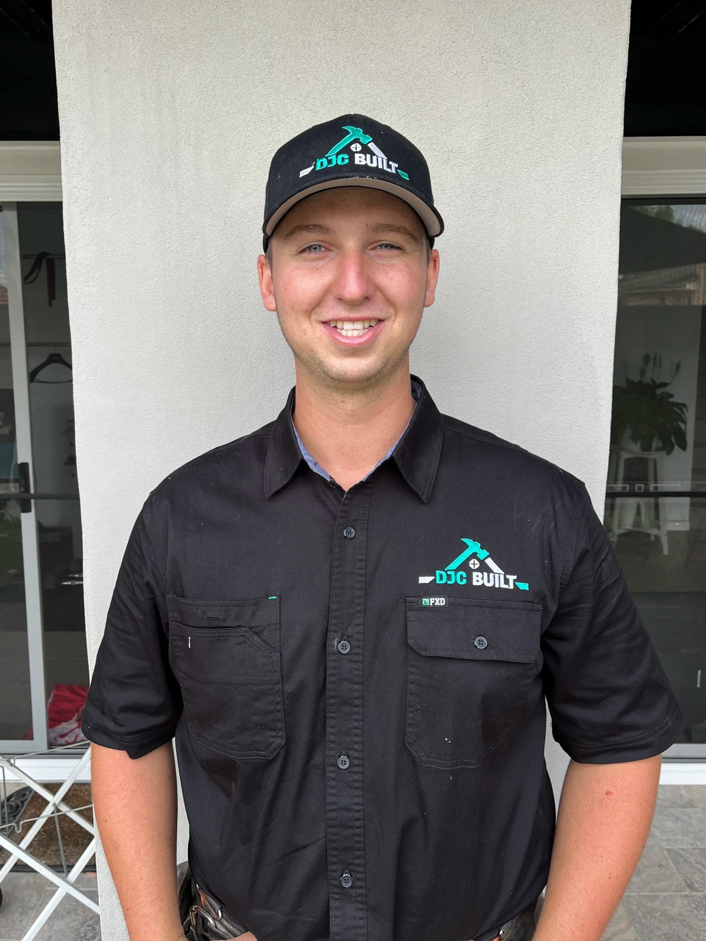 Young man smiling, wearing a black shirt and cap with 'DJC Built' logo, standing in front of a white wall.