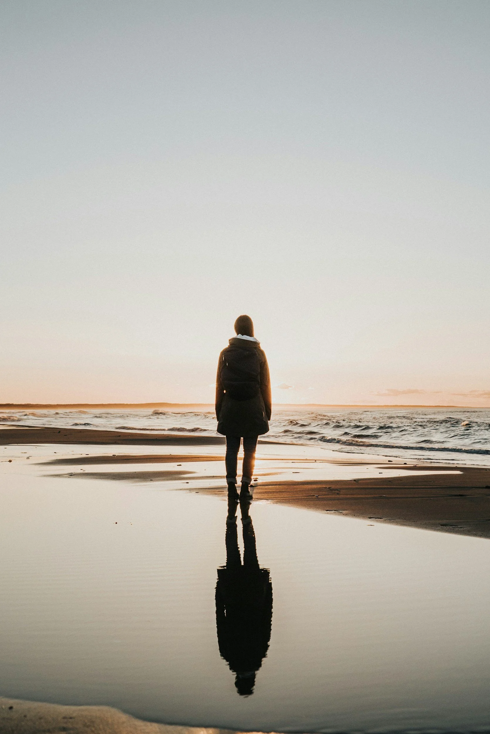 Person standing on beach near water at sunset with reflection visible on wet sand.