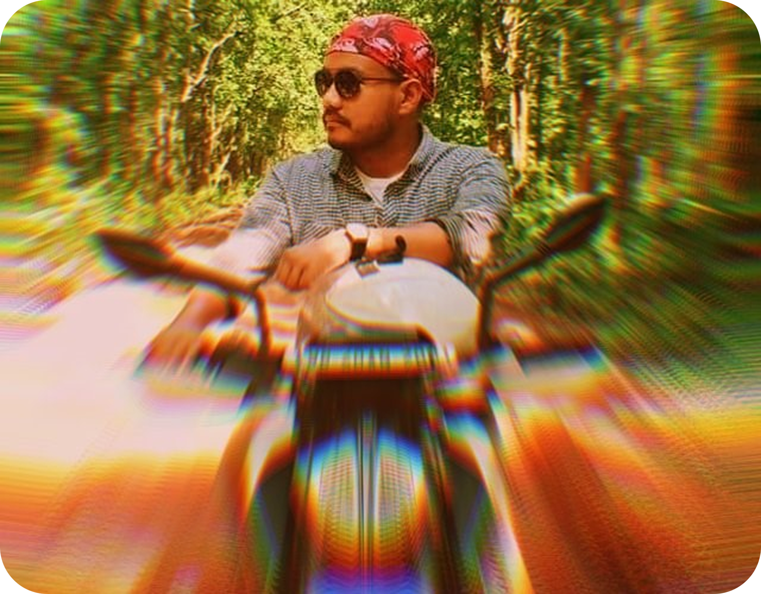 A man riding a motorcycle through a wooded area during daytime. He is wearing sunglasses, a red bandana, a checkered shirt, and a watch.