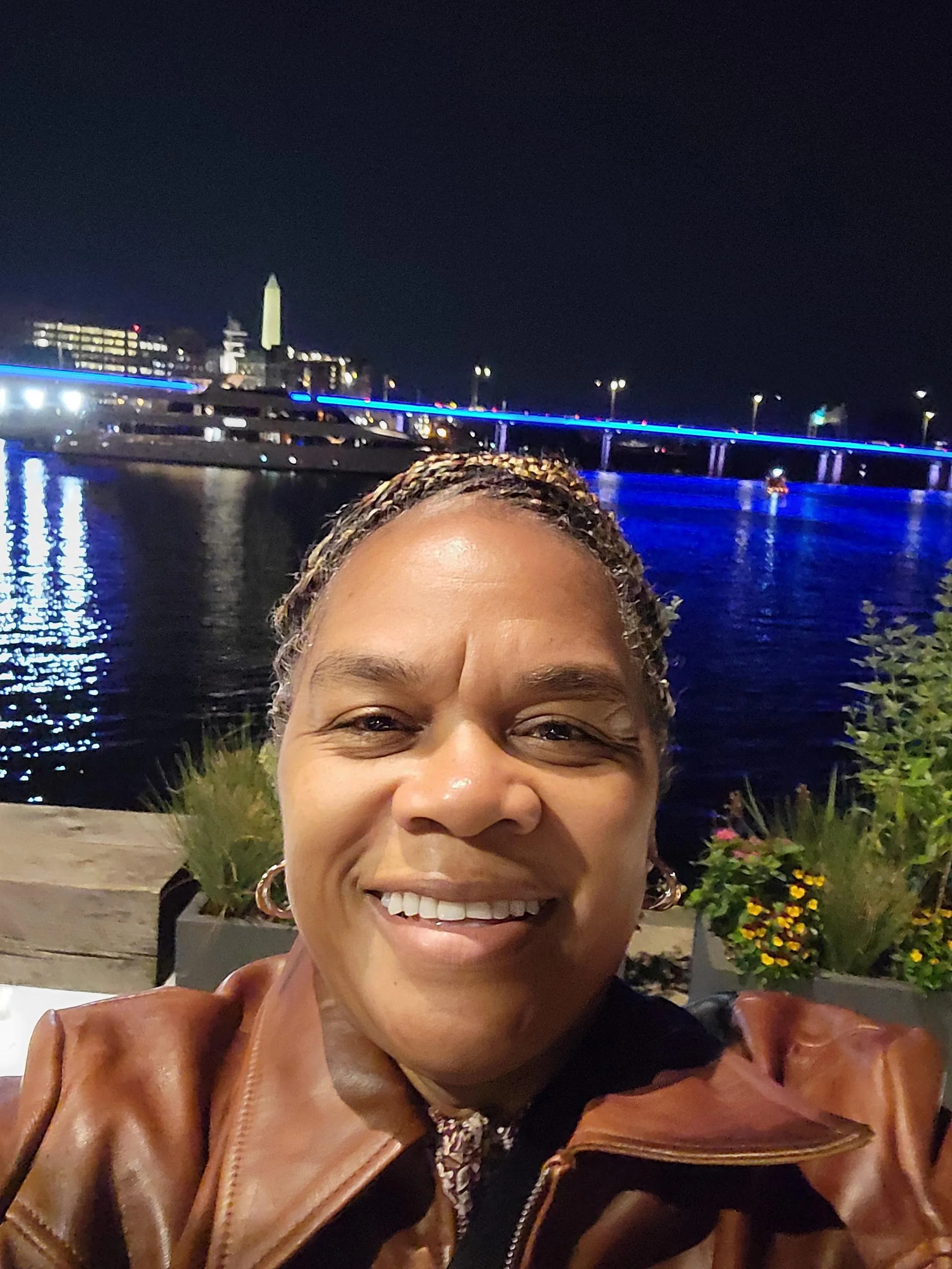 A woman taking a selfie at night near a body of water with a bridge illuminated in blue lights and city buildings in the background.