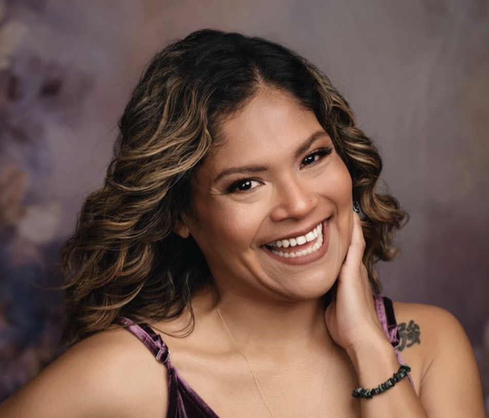 A woman with shoulder-length, wavy brown hair with blonde highlights, smiling and touching her neck, wearing a purple velvet top and jewelry, against a blurred background.