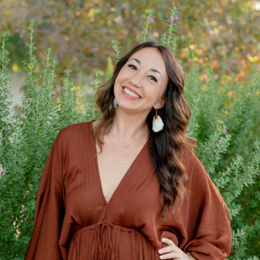 A woman smiling outdoors in front of green foliage, wearing a rust-colored dress, white tassel earrings, and layered necklaces.