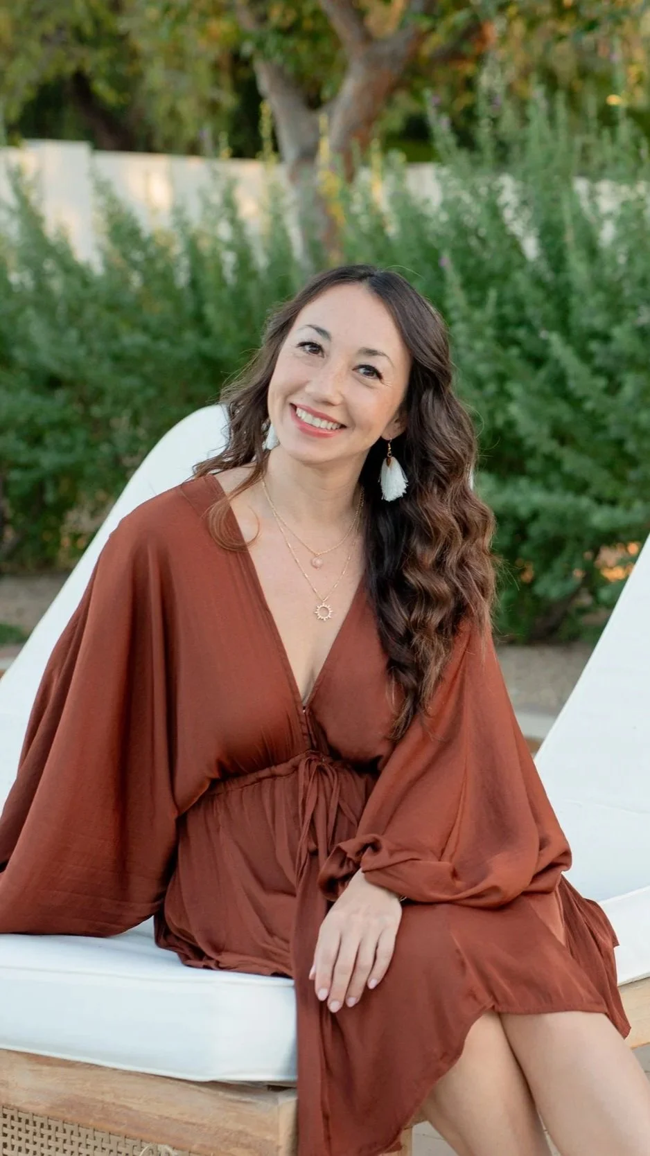 A woman with long, wavy brown hair sitting outdoors on a white cushioned bench, smiling at the camera, wearing a rust-colored dress and white tassel earrings, with green bushes and a tree in the background.