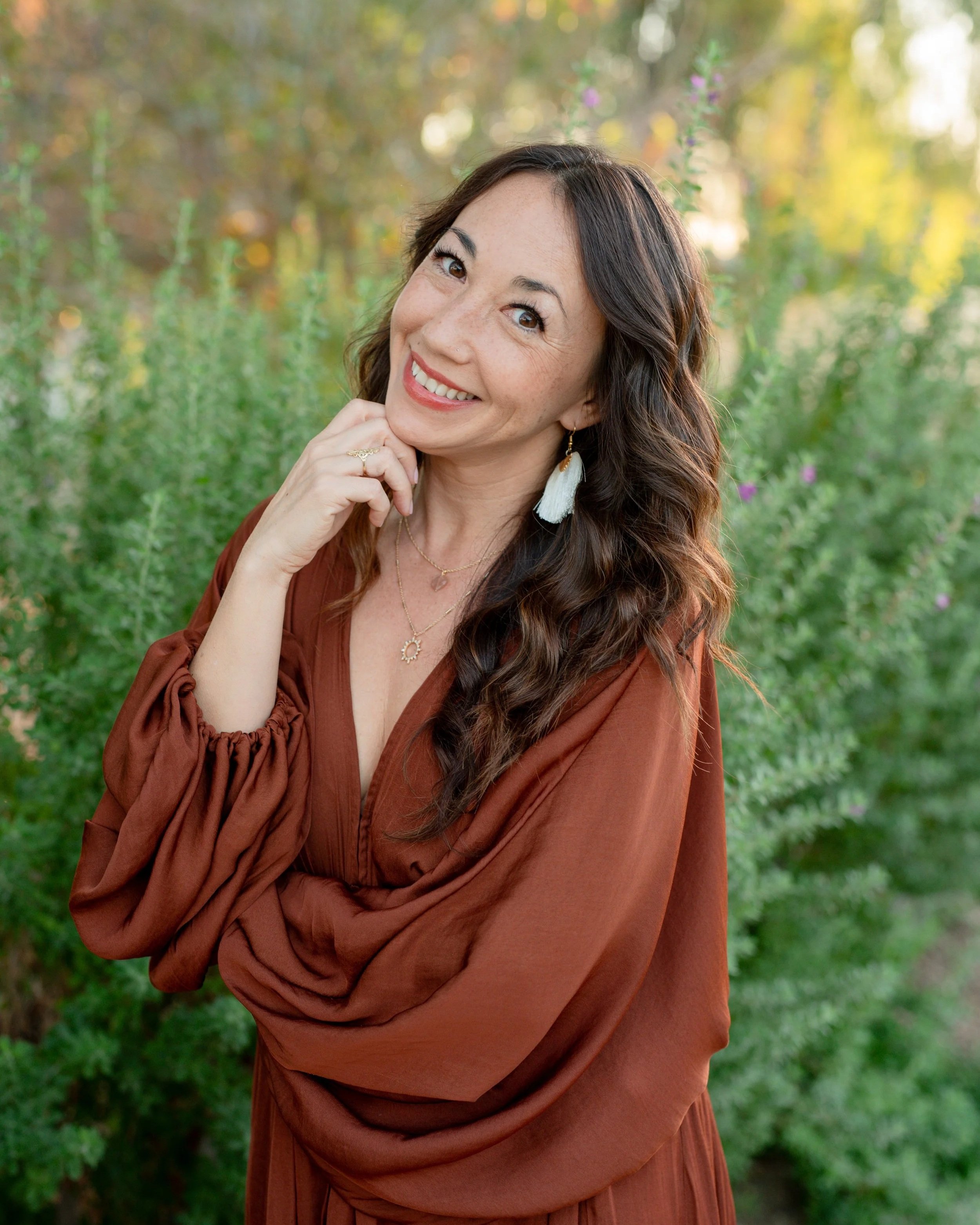A woman with long wavy hair, wearing a rust-colored dress and earrings, smiling outdoors with greenery and trees in the background.