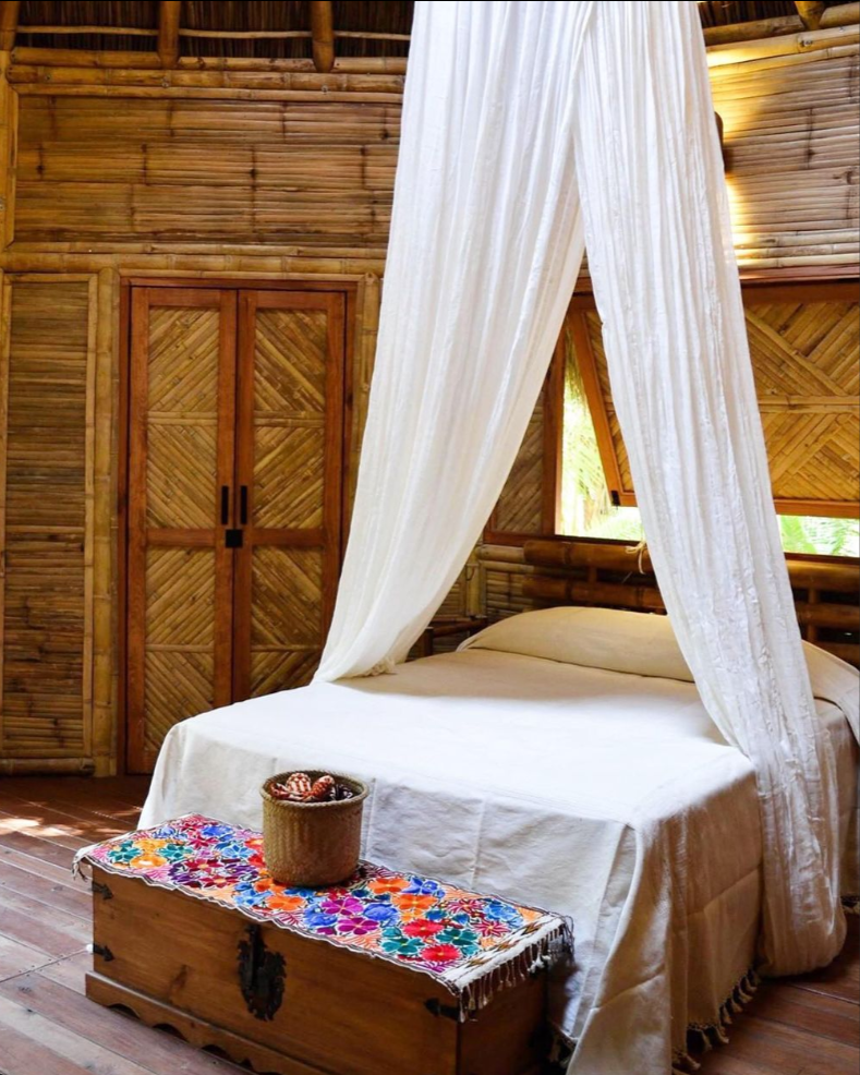 A rustic bedroom with a four-poster bed draped in white fabric, wooden walls, a chest with colorful embroidery, and a small window with natural light.