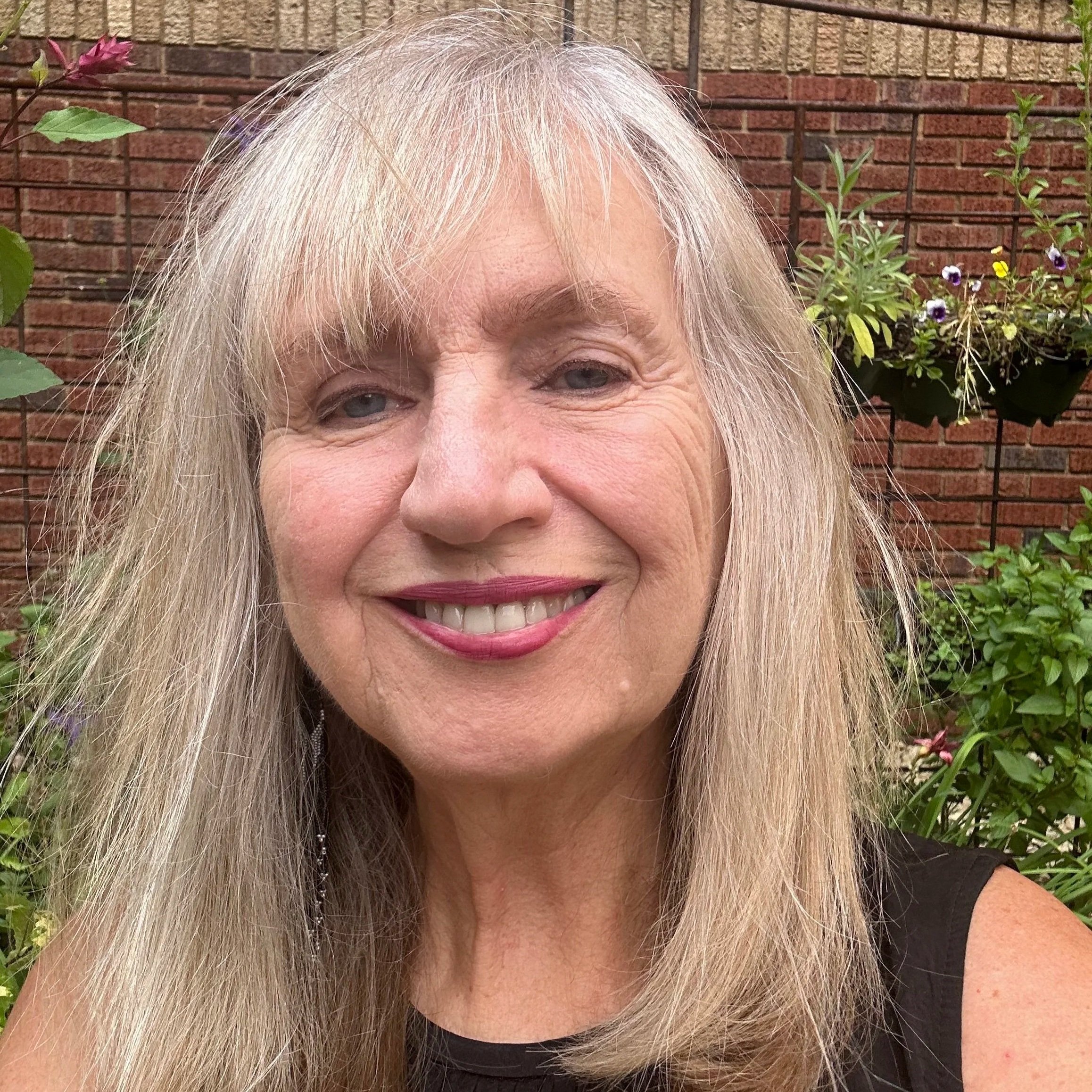 Close-up of a smiling woman with long blonde hair and blue eyes, standing outdoors with a brick wall and hanging flowers in the background.