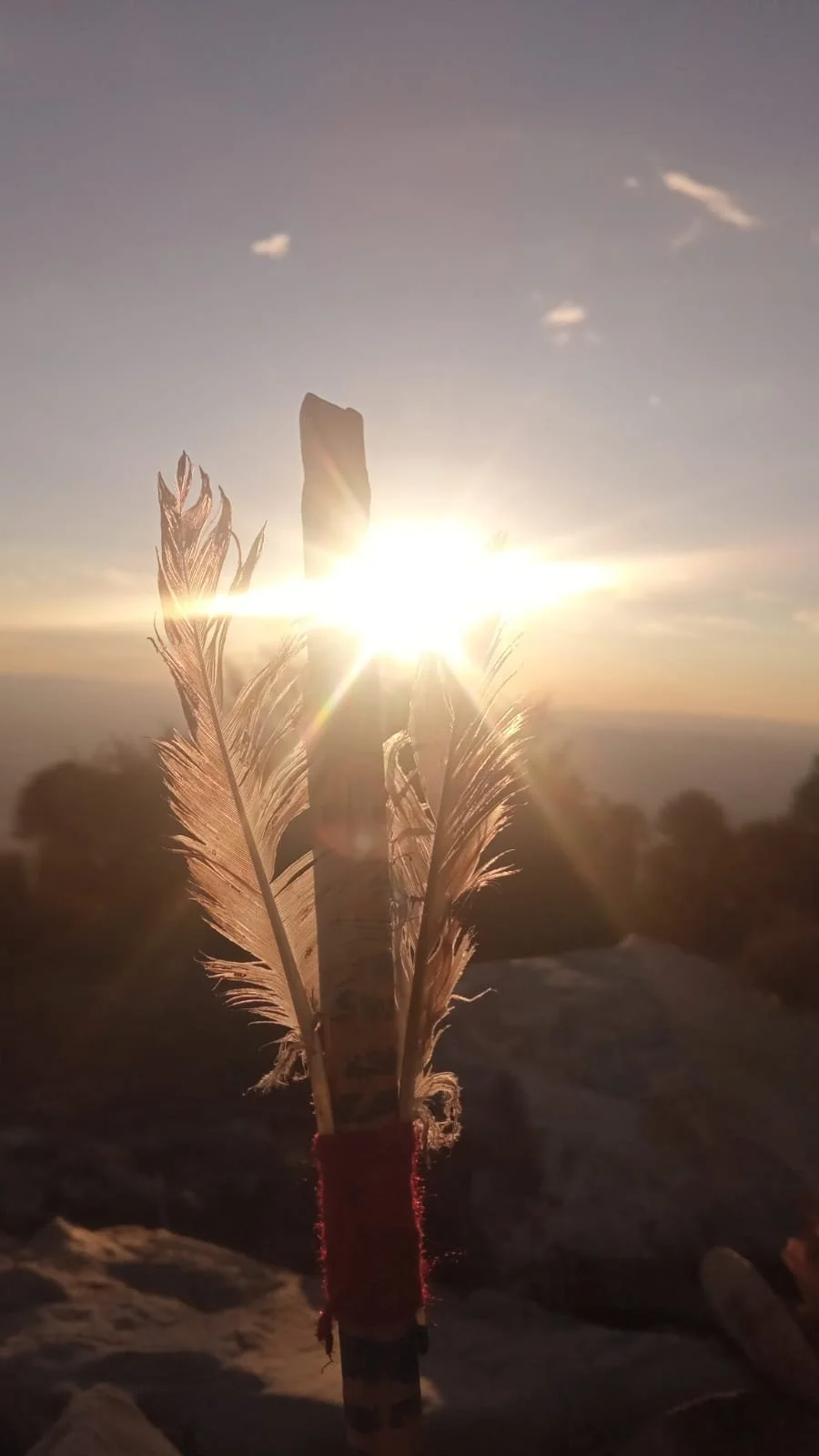 A wooden stick with feathers attached, wrapped in red fabric, standing upright on a rocky surface during sunset with the sun shining behind it, creating a lens flare and silhouettes of trees in the background.