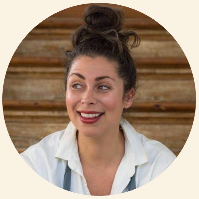 A woman with dark hair in a bun, smiling and looking to the side, wearing a white shirt in front of a wooden background.