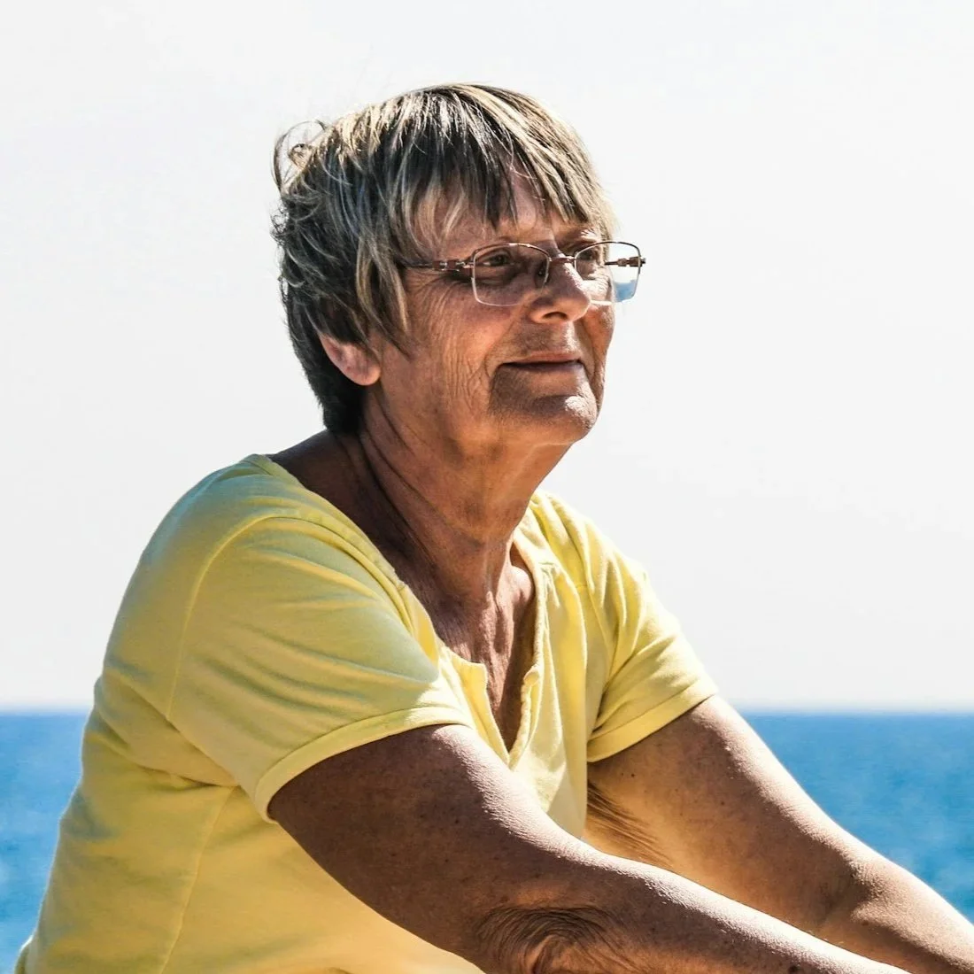 An elderly woman with short gray hair and glasses, wearing a yellow shirt, looking pensively over the ocean.