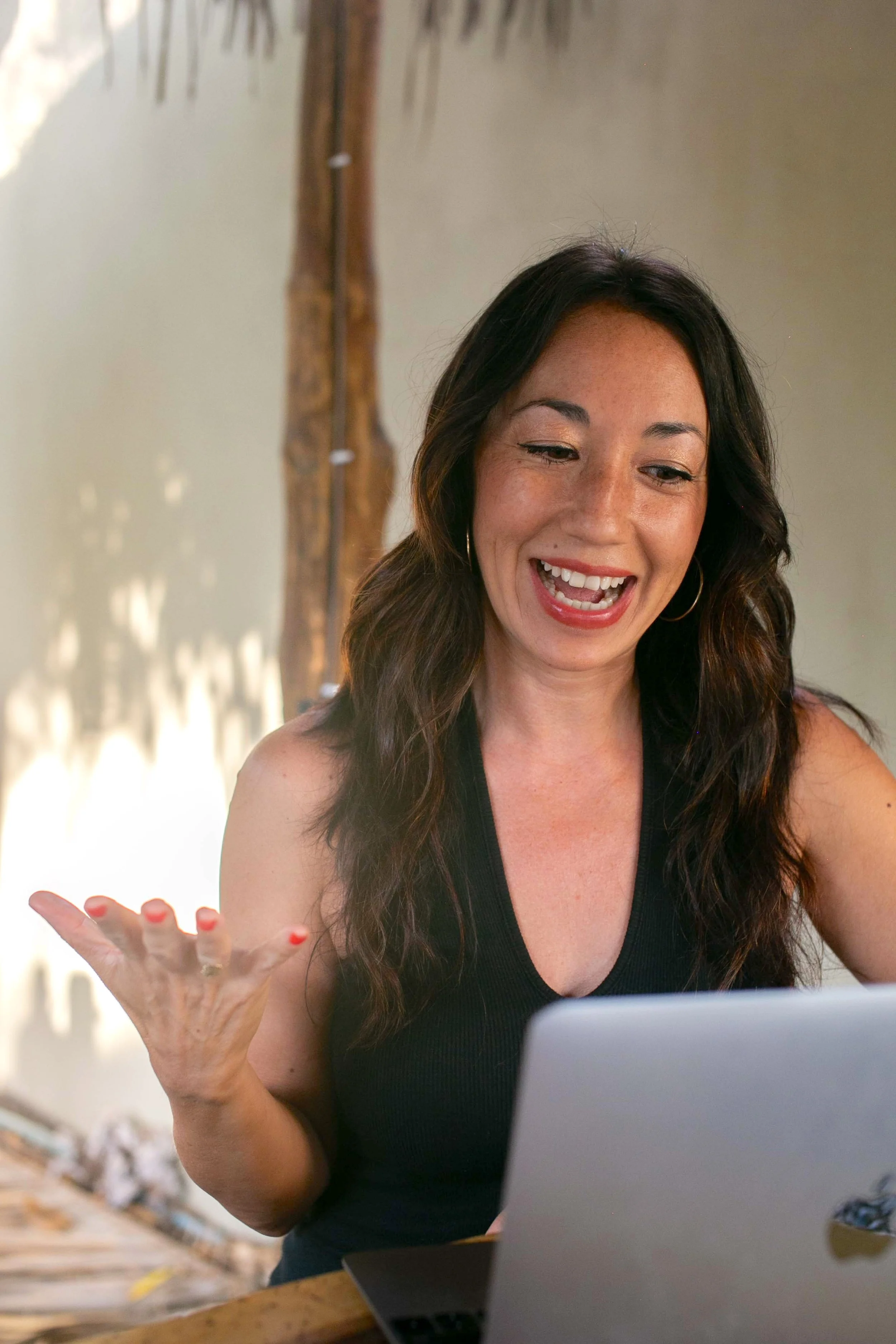 A woman with long dark hair, wearing a black top, is smiling and gesturing with her hand while looking at a laptop.