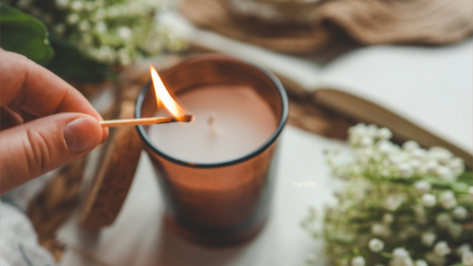 Person lighting a candle with a matchstick in front of a brown glass candle holder, surrounded by white flowers.