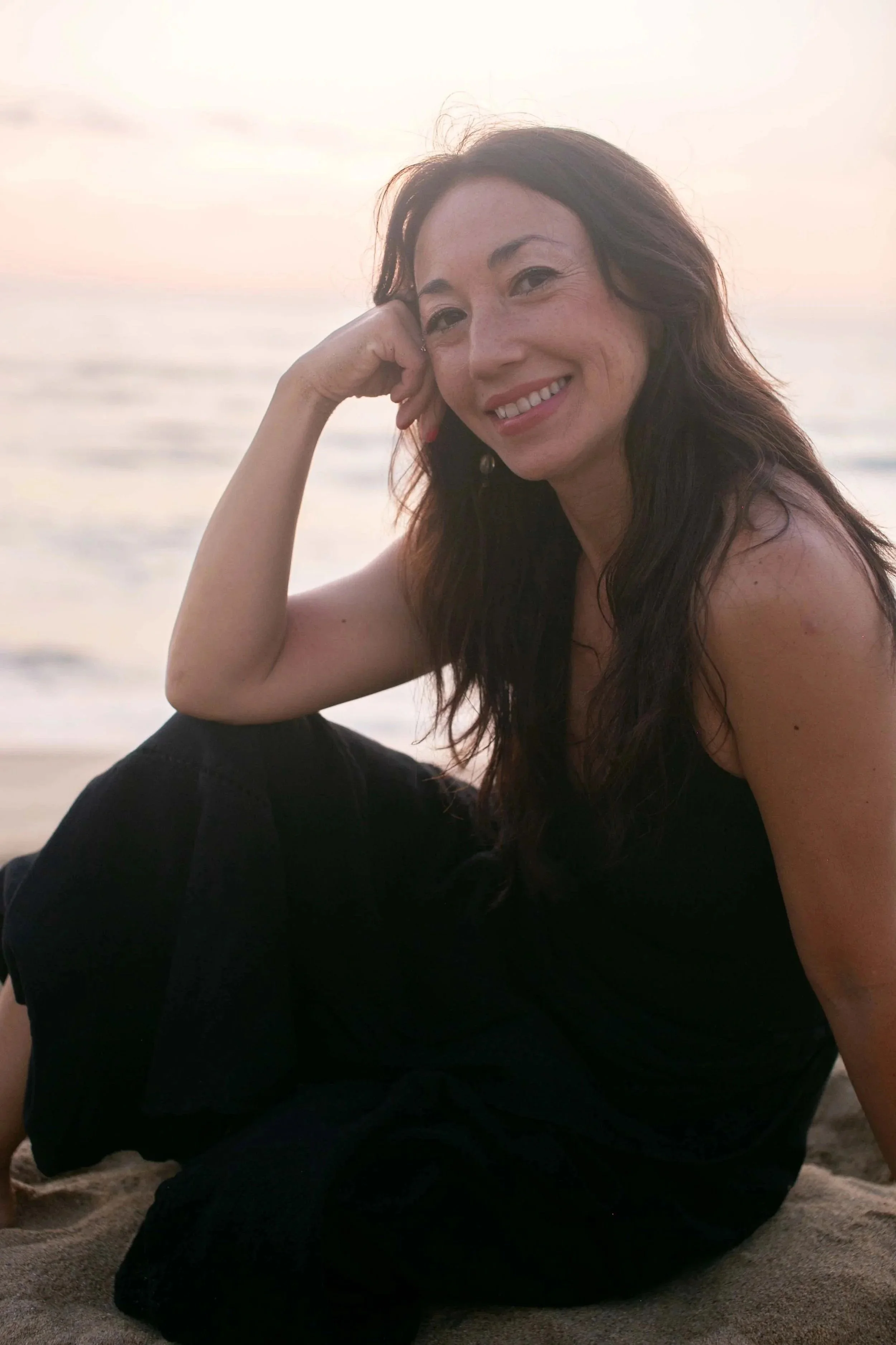 A woman with long dark hair and a black dress sitting on the beach at sunset, smiling at the camera.