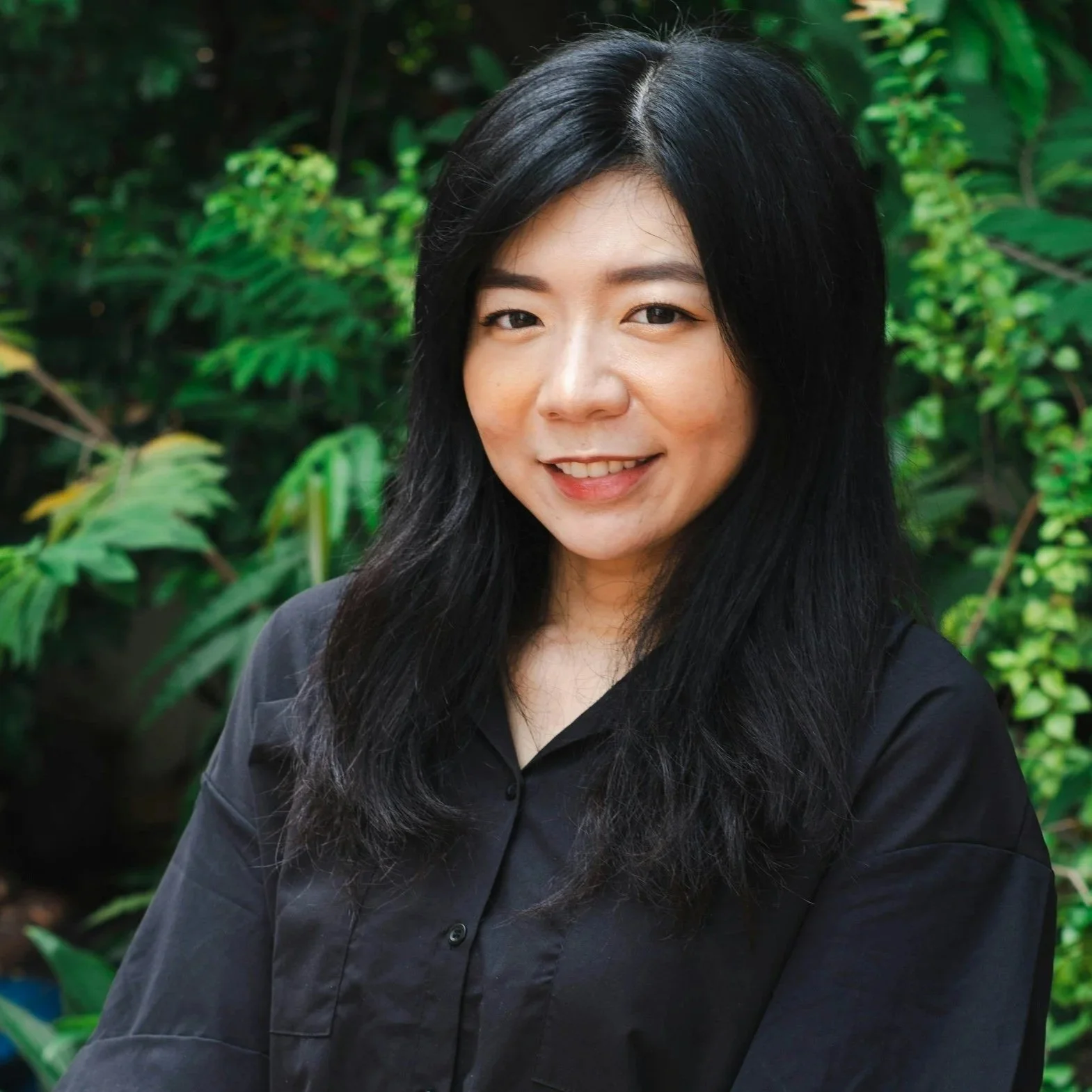 A woman with long black hair smiling outdoors with green foliage in the background.