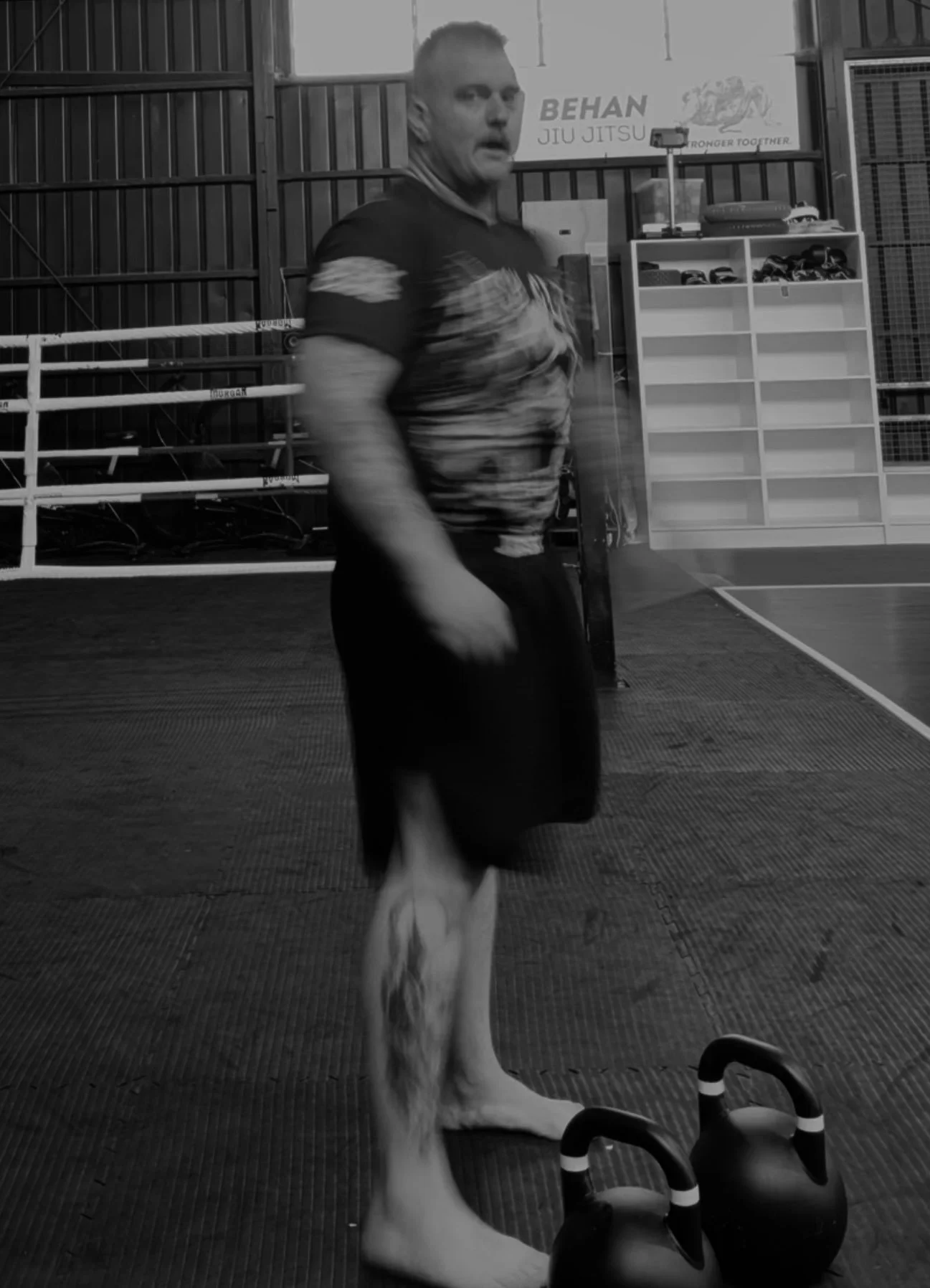 A man with tattoos on his leg, wearing a t-shirt and shorts, standing barefoot in a gym with kettlebells on the floor in front of him.