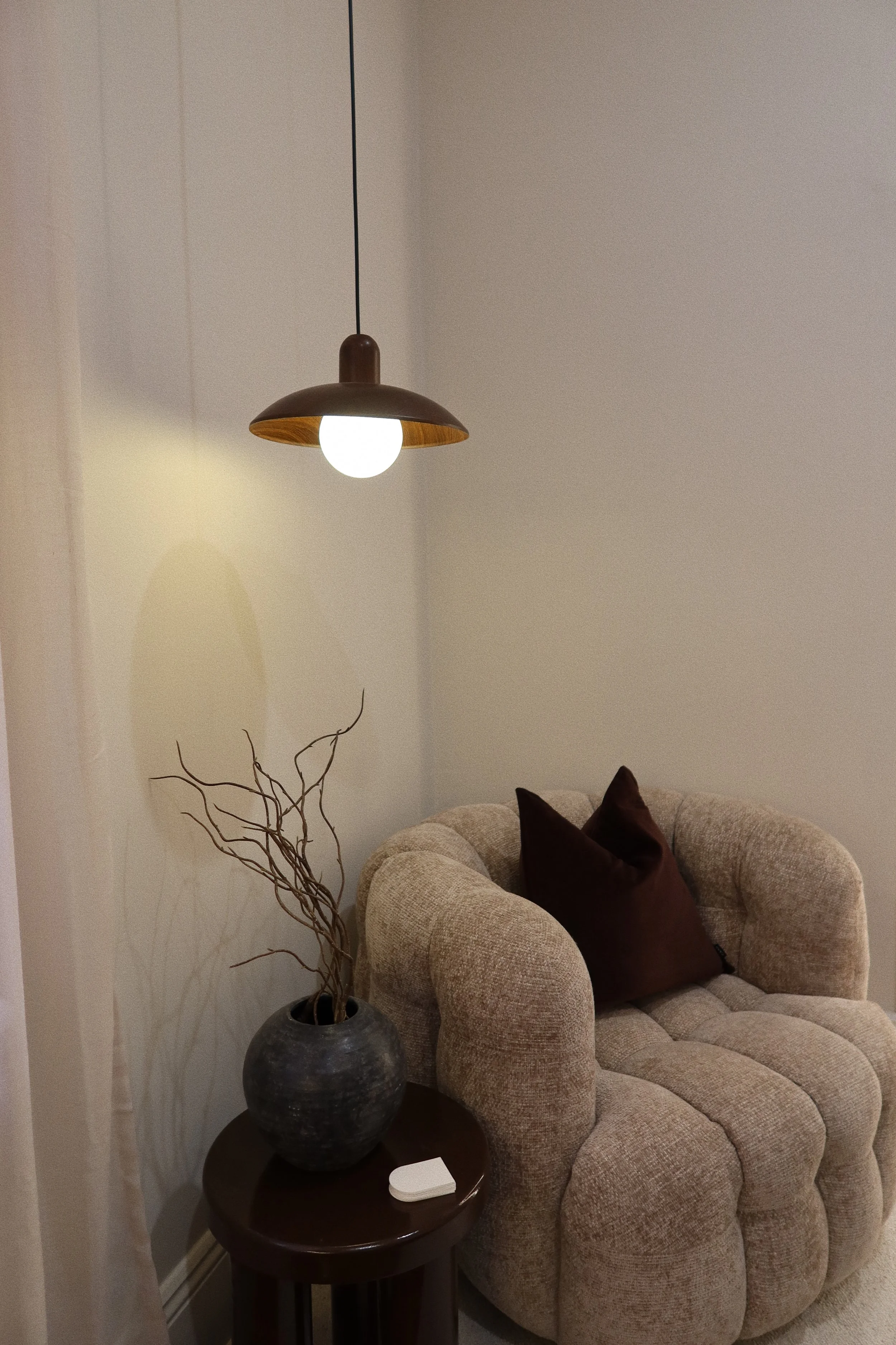 A cozy beige armchair with a dark brown pillow, next to a small round wooden table holding a large black vase with twigs and a white object, illuminated by a modern pendant light hanging from the ceiling.